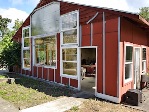 Exterior view of a red building with white trim and multiple windows. The building has an open door revealing a glimpse of the interior with some furniture. There is a small concrete path leading to the entrance and some greenery around the building.