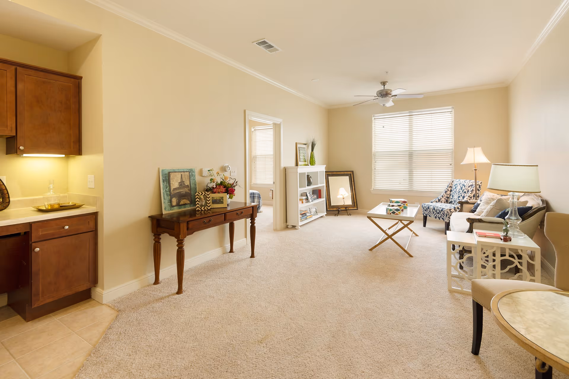 A bright living room with beige walls and carpet, featuring a white sofa, patterned armchair, coffee table, side tables with lamps, a small bookshelf, and a wooden console table with decorative items. A ceiling fan and a large window with blinds provide light to the room. Part of a kitchen with wooden cabinets is visible on the left.