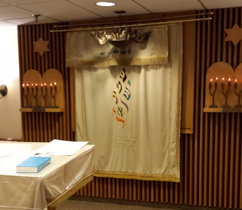 Interior view of a small prayer or meditation room with wooden slatted walls. A cream-colored curtain with Hebrew text and decorative embroidery hangs in the center. On either side of the curtain, there are wooden menorah-shaped candle holders with lit candles. A table covered with a white cloth and a blue book is positioned in the foreground.
