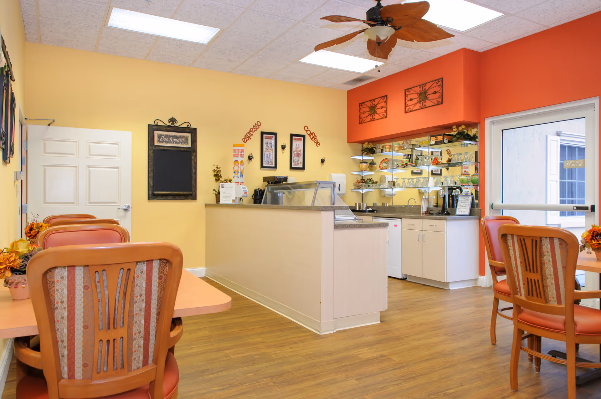 A cozy dining area with wooden chairs and tables, a counter with a glass display case, and a small kitchen area with shelves holding cups and plates. The walls are painted yellow and orange, and there is a ceiling fan with wooden blades. A door with a window leads outside.