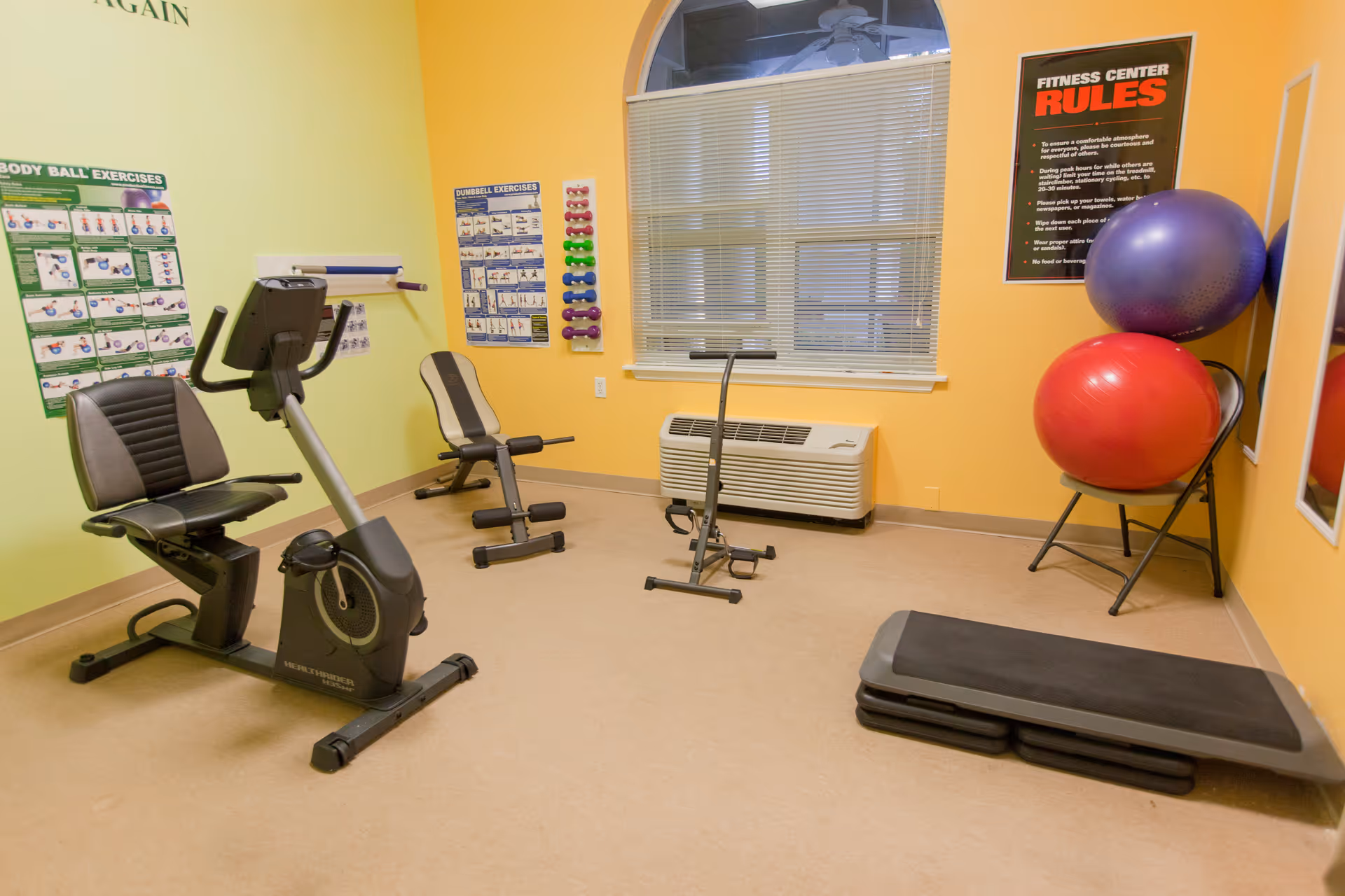 A small fitness center room with exercise equipment including a recumbent bike, a weight bench, an ab exercise machine, two large exercise balls on a chair, and a step platform. The walls are painted yellow and green, with exercise instruction posters and fitness center rules displayed. A window with blinds is centered on the back wall.