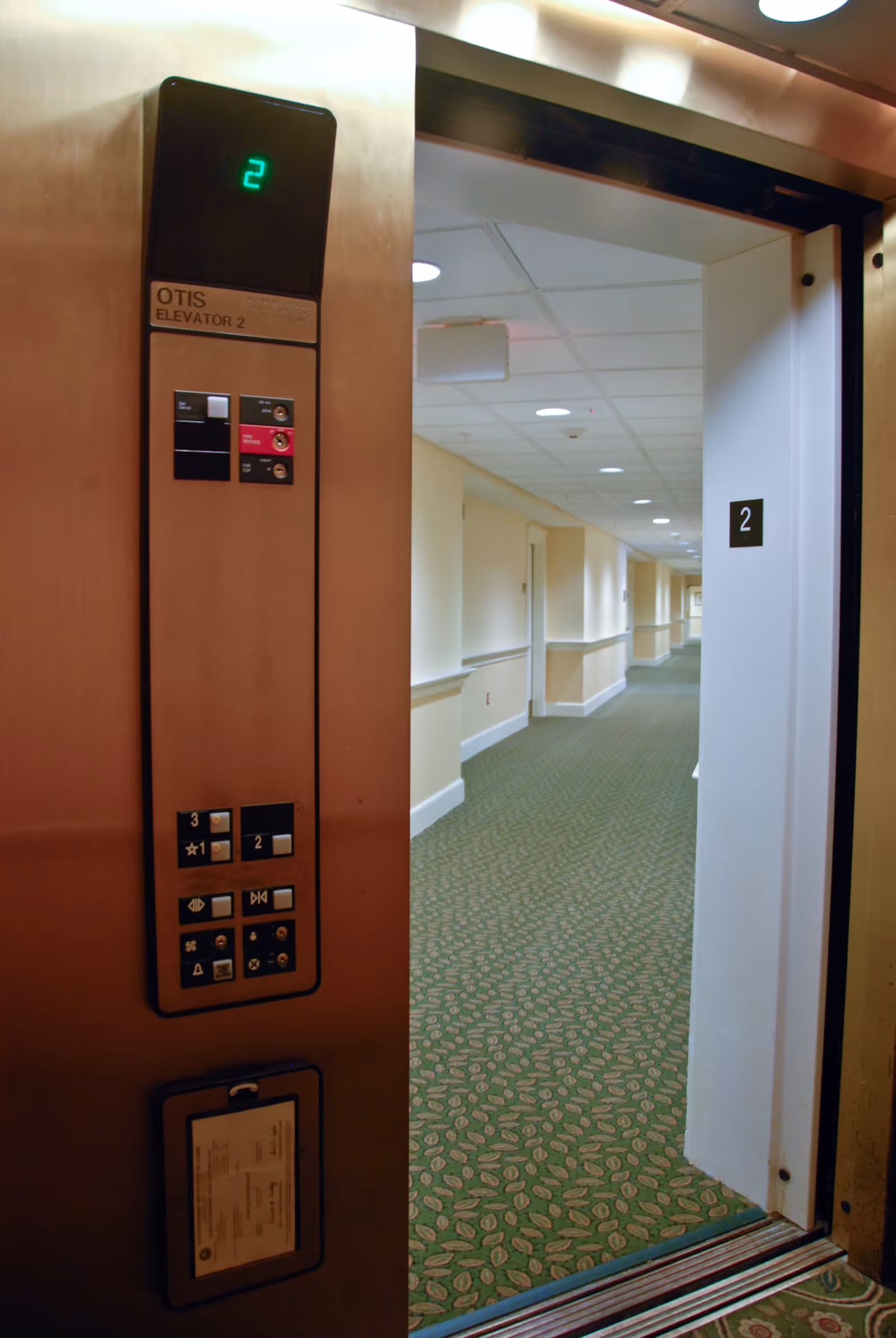 Open elevator door showing the interior hallway of a senior living facility with green patterned carpet and beige walls. The elevator panel displays the number 2 and has various buttons and indicators.