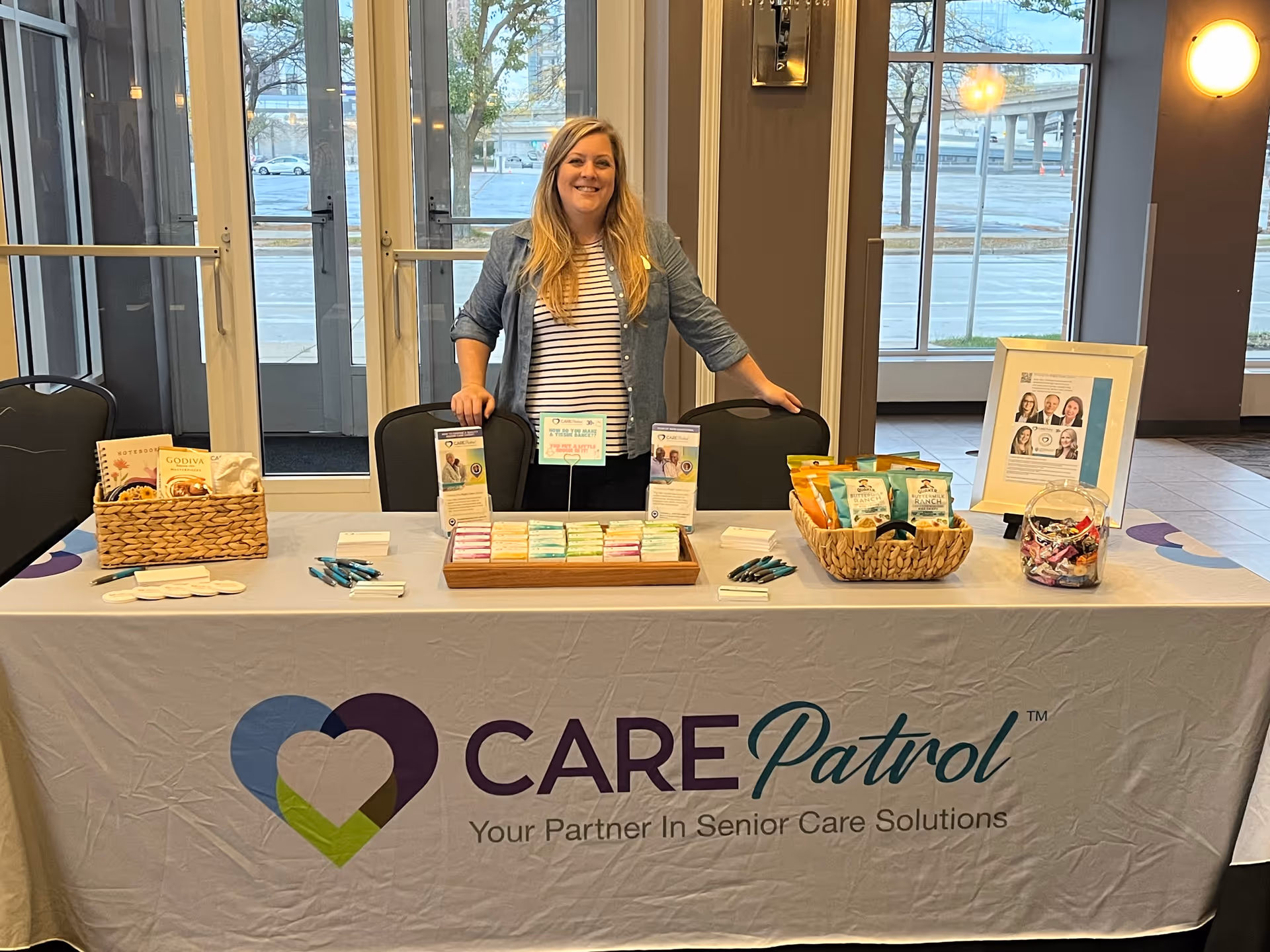A woman standing behind a table covered with a CarePatrol branded tablecloth. The table displays brochures, snacks, pens, and promotional materials. The setting appears to be an indoor event space with large windows and glass doors in the background.