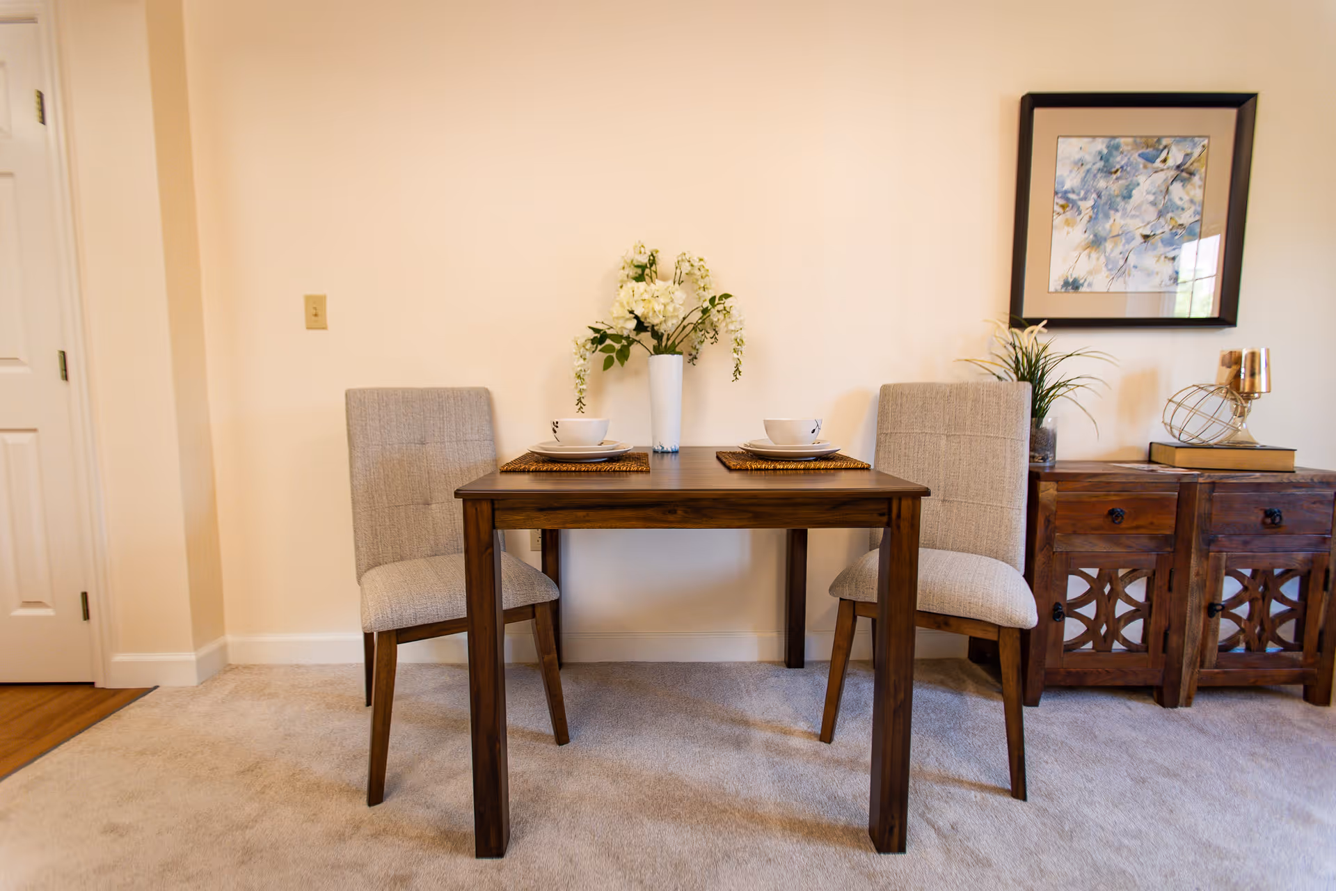 A small dining area with a wooden table set for two, featuring two beige upholstered chairs. On the table, there are two place settings with white bowls and plates on woven placemats, and a white vase with white flowers. To the right, there is a wooden sideboard with decorative items including a plant, a gold candle holder, and a framed abstract painting on the wall above it. The room has beige walls and carpeted flooring.