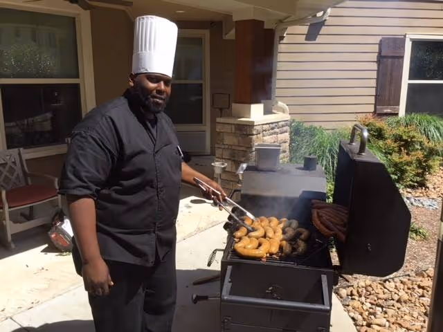 A chef wearing a white chef hat and black uniform is grilling sausages on a barbecue grill outside a building with beige siding and stone accents. There is a chair and a window in the background, and some greenery to the right.