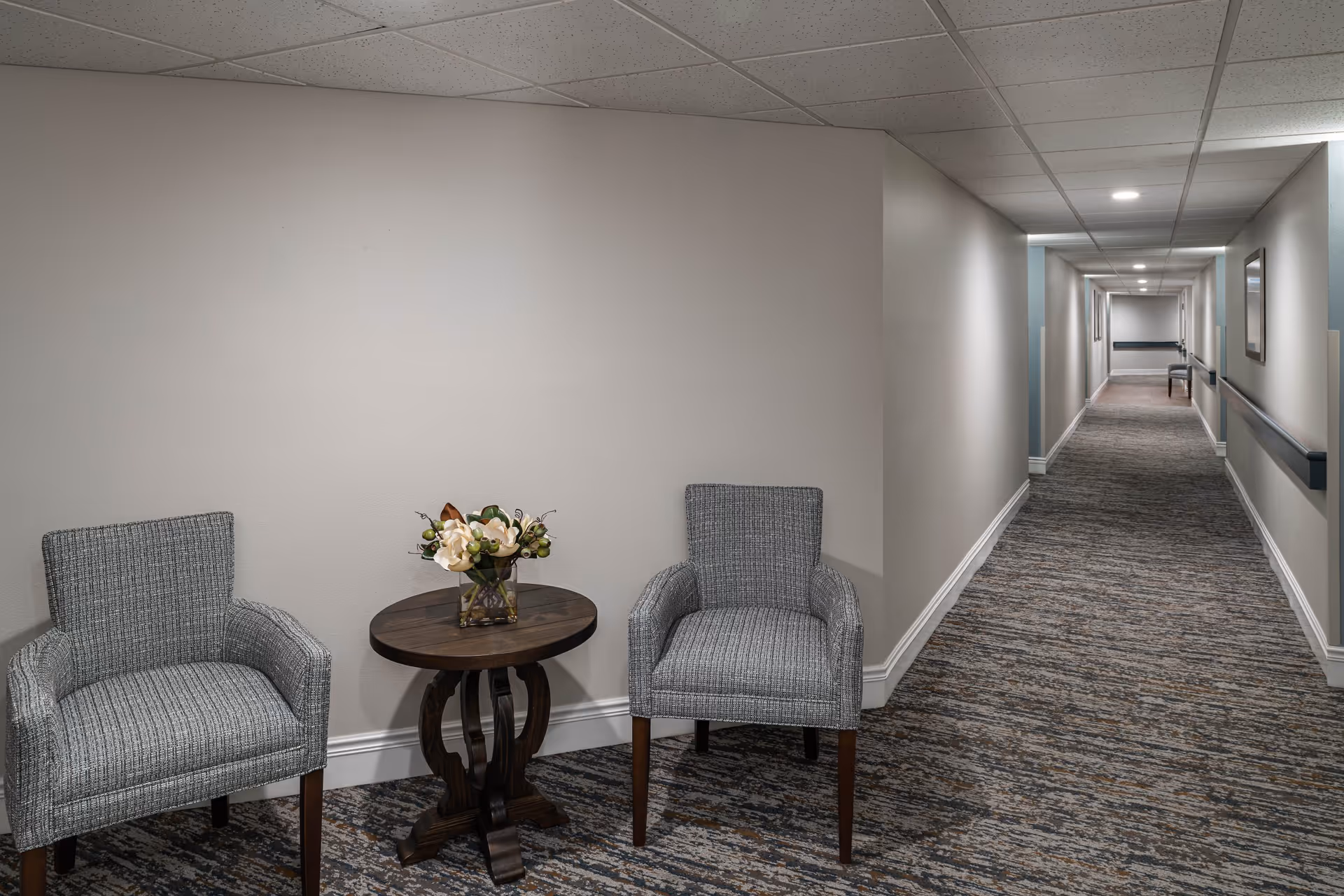 A long, carpeted hallway in a senior living facility with two gray upholstered chairs and a small round wooden table with a floral arrangement placed against a beige wall on the left side. The hallway has handrails on both sides and framed pictures on the walls, with ceiling lights illuminating the space.
