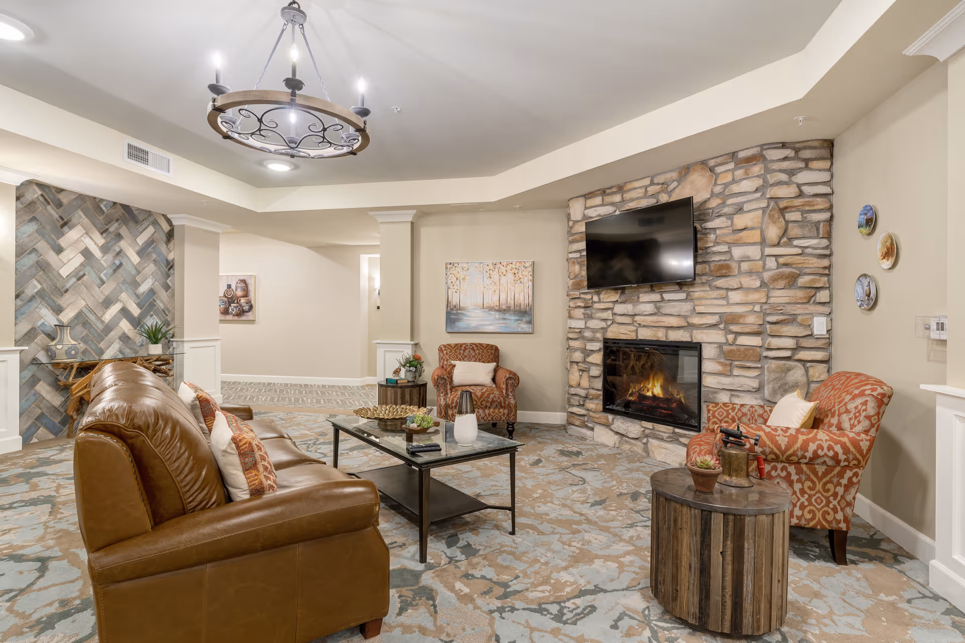 A cozy senior living facility common area with a brown leather sofa, two patterned armchairs, a glass coffee table, and a stone fireplace with a mounted flat-screen TV above it. The room features a decorative chandelier, patterned carpet, and artwork on the walls.