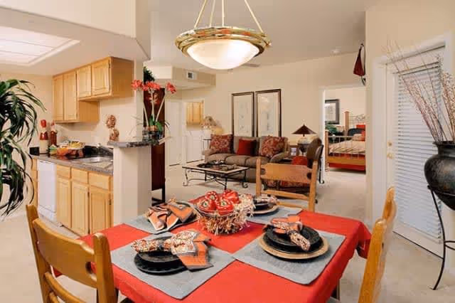 View of a dining area with a table set for four with black plates, patterned napkins, and a red tablecloth. Behind the dining area is a kitchen with wooden cabinets and a granite countertop. Further back is a living room with a sofa, armchair, coffee table, and framed artwork on the wall. A bedroom is visible through an open doorway to the right.