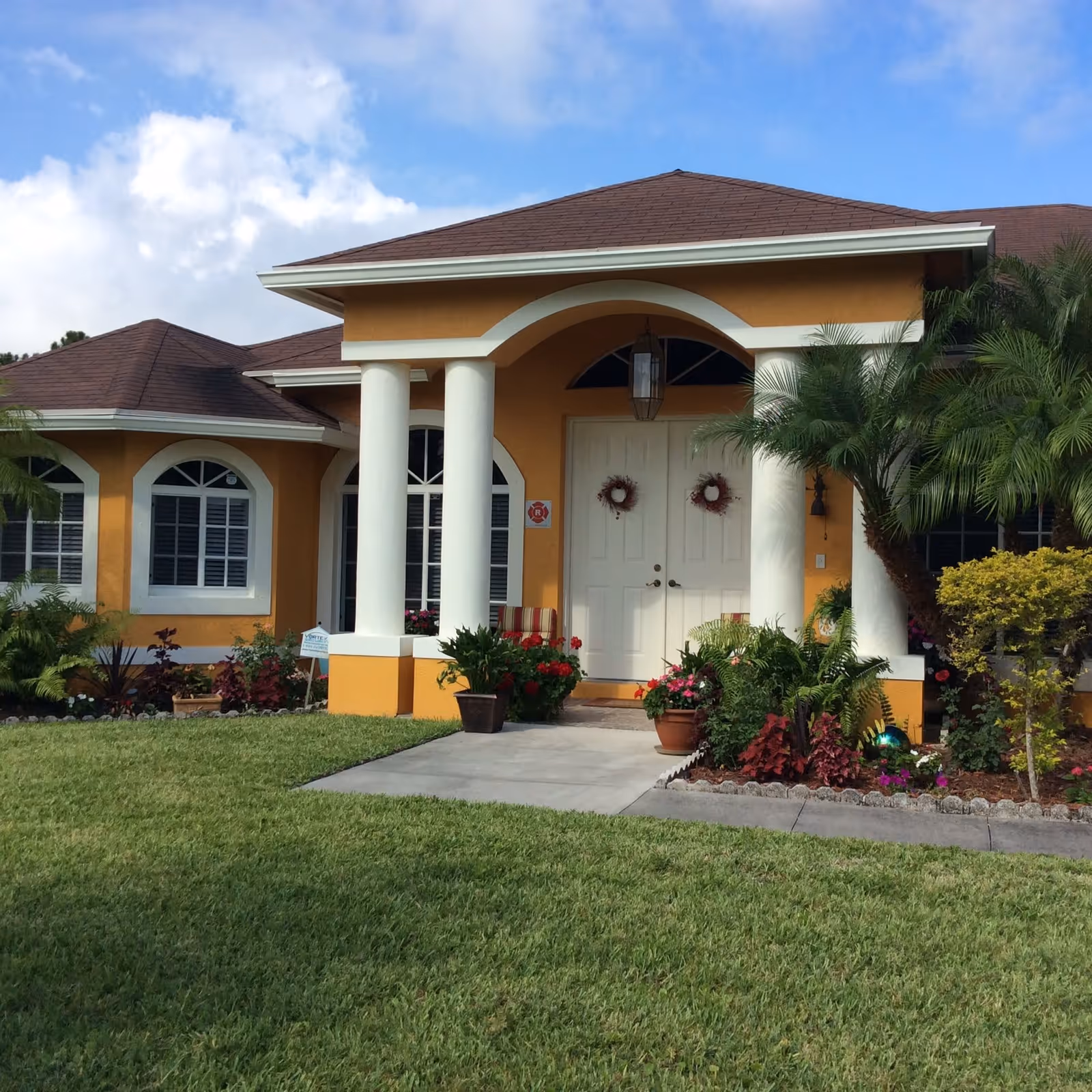 Front exterior view of a single-story building with a yellow-orange facade, white double doors decorated with wreaths, white columns supporting a small porch roof, and well-maintained landscaping including green grass, shrubs, and potted plants.