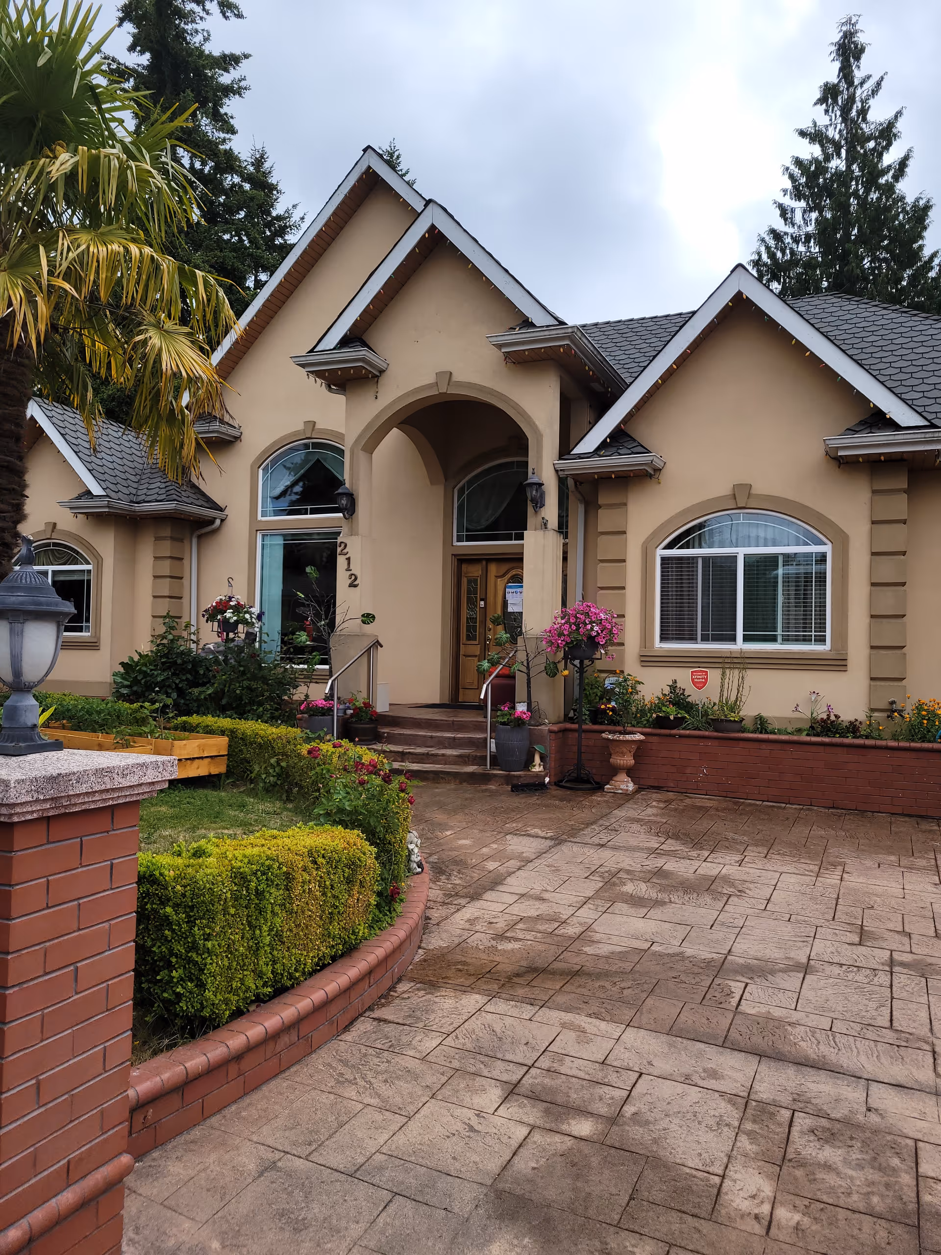 Front exterior view of a beige residential building with a peaked roof, arched doorway, and large windows. The entrance is accessed by a few steps with handrails on both sides. There are well-maintained shrubs, flowers, and a palm tree in the landscaped garden area along the walkway leading to the door. The sky is cloudy.