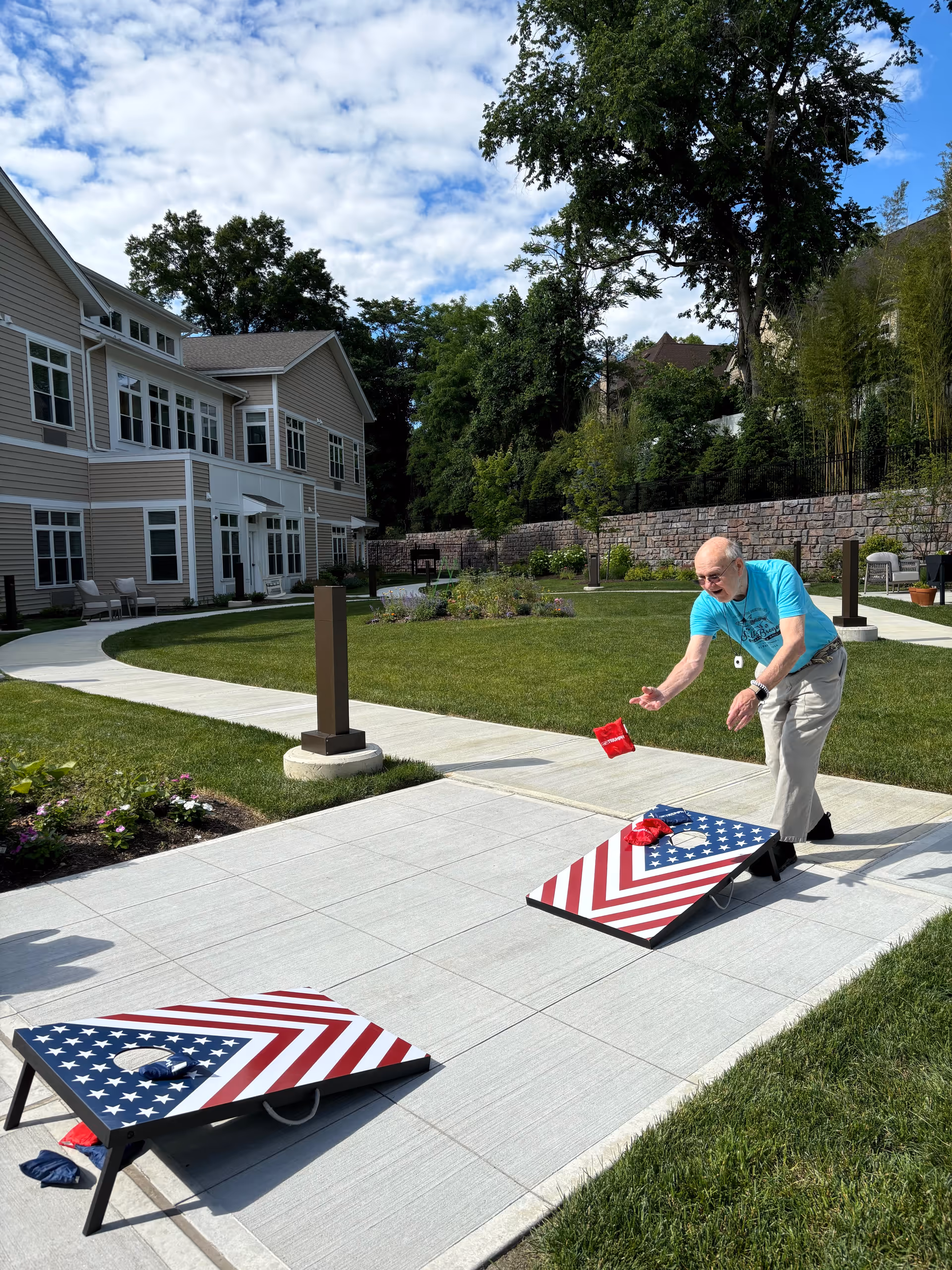 A person tossing a beanbag at an American-flag cornhole board on a paved courtyard in front of a two-story senior living building with lawn and trees.