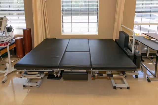 An adjustable black padded therapy table in a rehabilitation room with windows and medical equipment nearby.