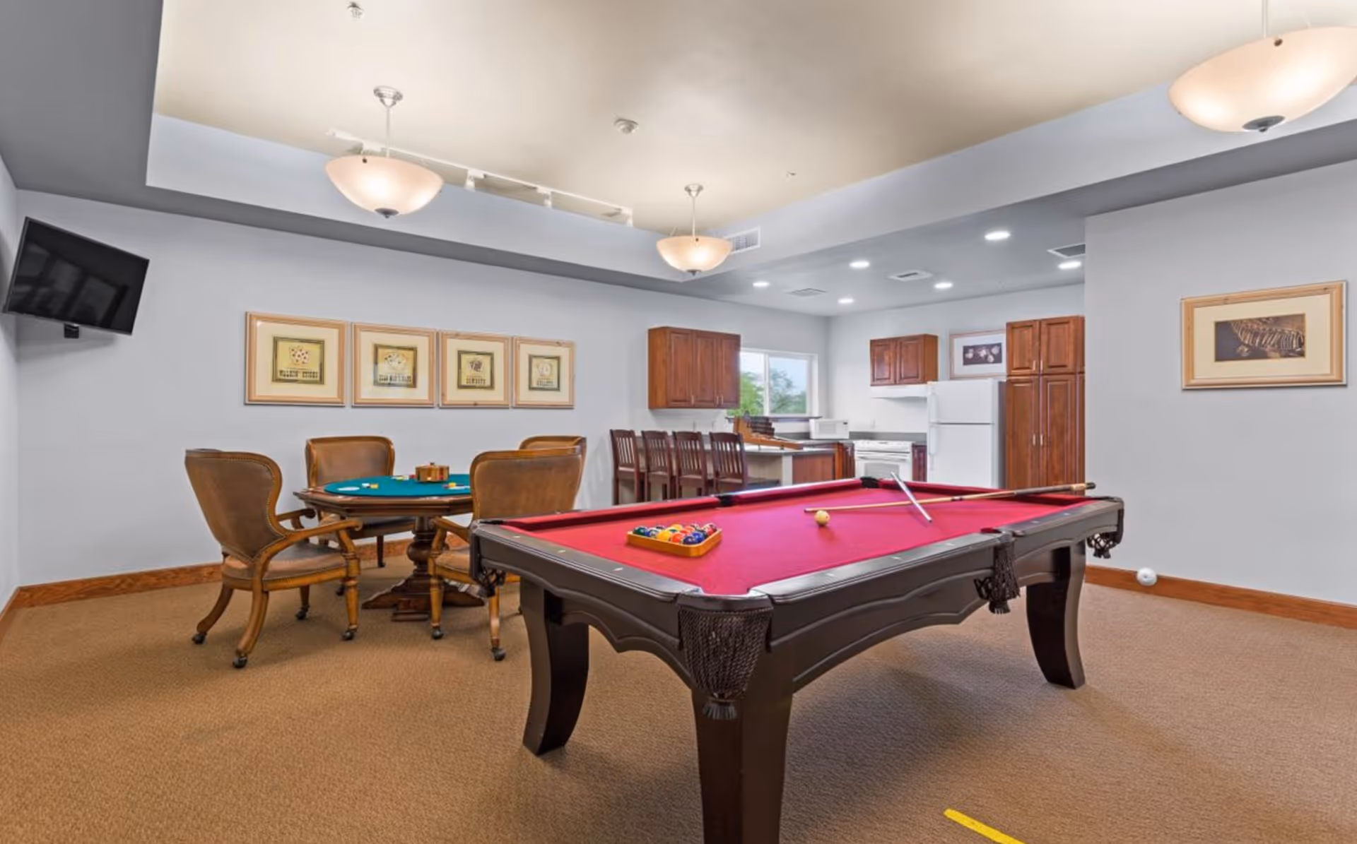 A recreational room featuring a red felt pool table with pool balls and a cue stick, a round card table with four chairs, a wall-mounted TV, and a kitchen area with wooden cabinets, a refrigerator, and a stove in the background. The room has beige carpet, light gray walls, framed artwork, and ceiling light fixtures.