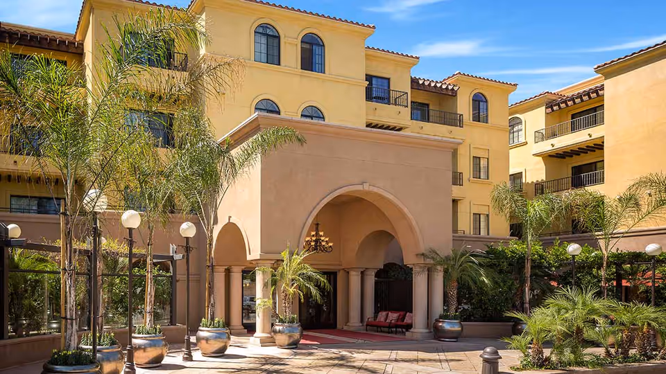 Exterior view of a multi-story senior living facility with yellow stucco walls, arched windows, and a covered entrance supported by columns. The courtyard features palm trees, potted plants, and outdoor seating under a clear blue sky.