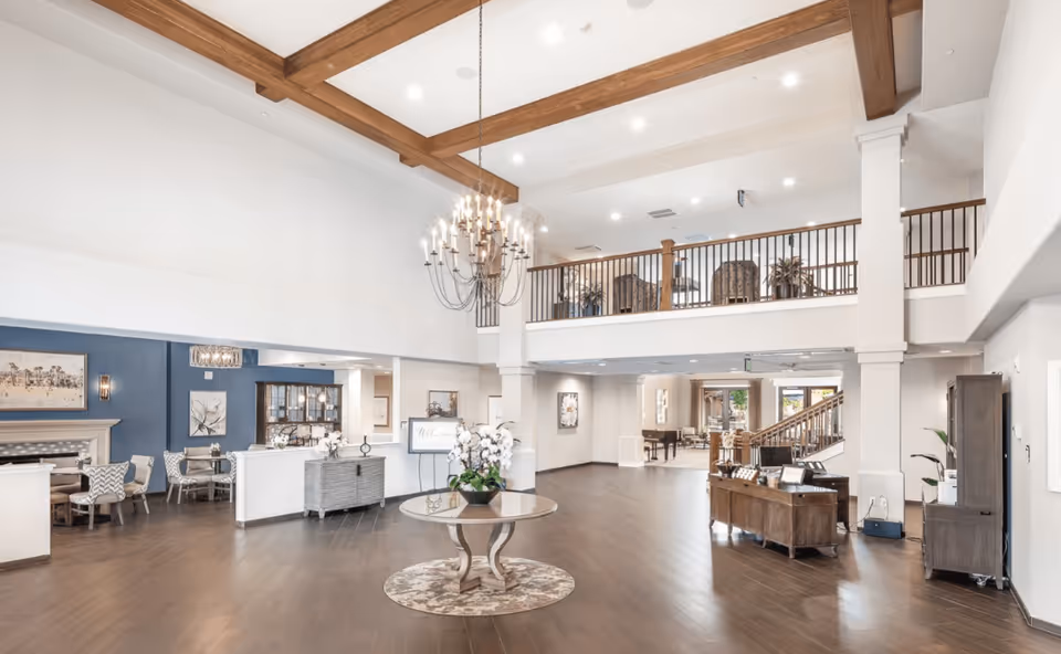 Spacious senior living facility lobby with high ceilings, wooden beams, a large chandelier, and a round table with a flower arrangement in the center. There are seating areas with chairs and tables along the walls, a staircase leading to an upper balcony with additional seating, and various pieces of furniture and decor throughout the space.