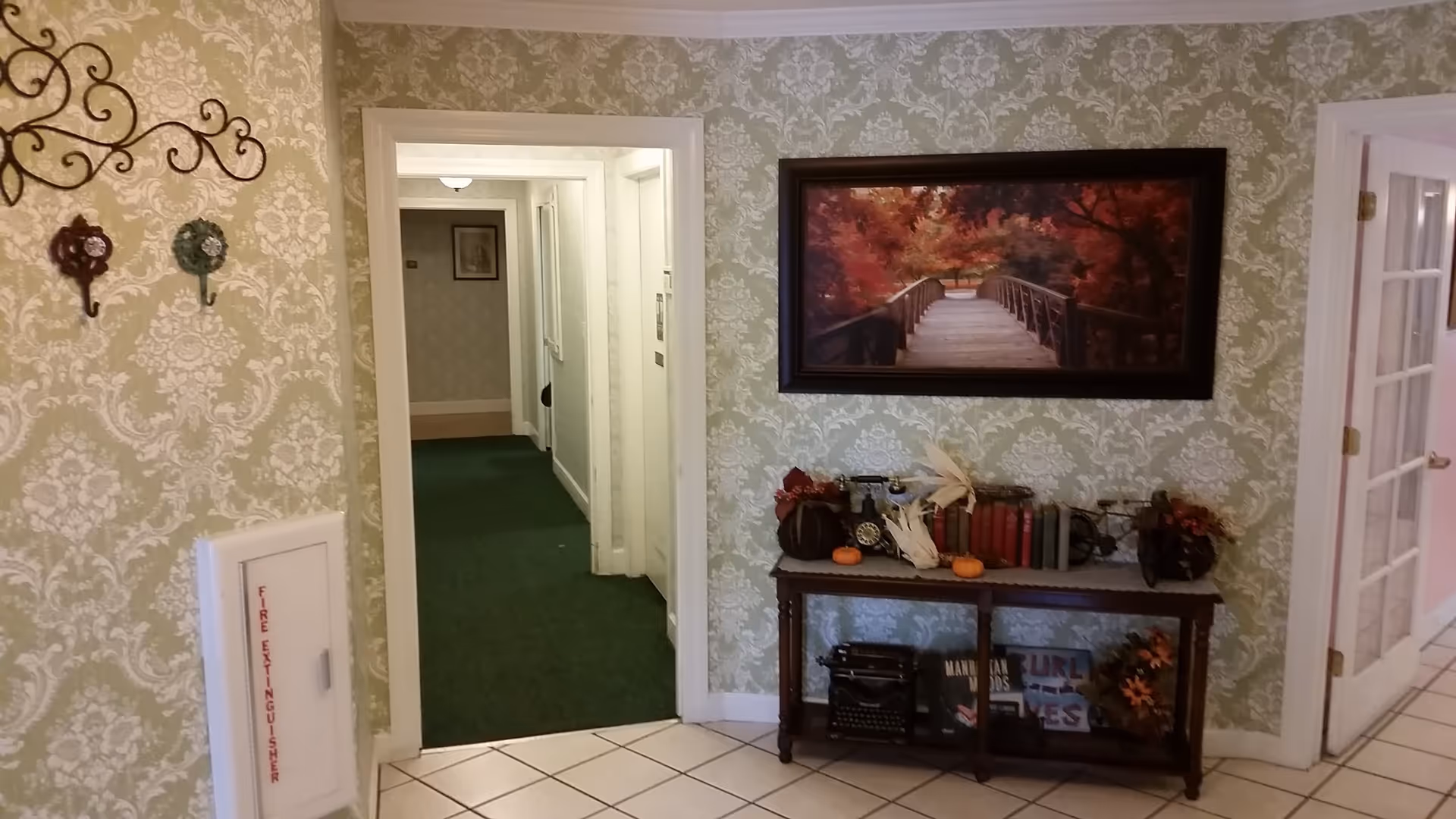 Interior hallway area with patterned wallpaper, a framed landscape painting above a decorated console table and a doorway leading to a carpeted corridor.