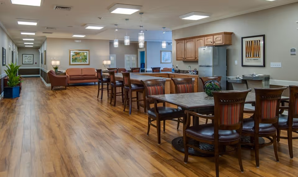 Interior view of a senior living facility common area featuring a kitchen with wooden cabinets and a refrigerator, a dining table with chairs, a seating area with sofas and armchairs, and a long hallway with framed artwork and plants.