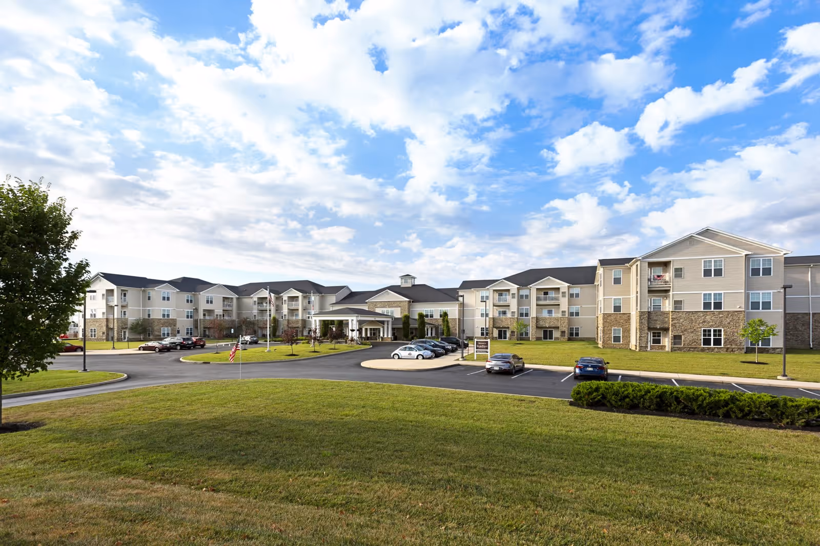 Exterior view of a large, multi-story senior living facility with beige siding and stone accents, surrounded by a well-maintained lawn and parking lot under a partly cloudy sky.