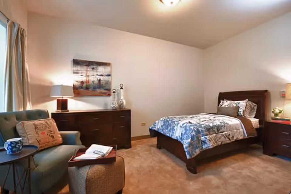 A cozy bedroom with a single bed featuring blue and white floral bedding, a wooden dresser with decorative items and a lamp, a comfortable armchair with a pillow, a small side table with a cup, and a soft ottoman. The room has beige carpet and neutral walls with a framed artwork above the dresser.