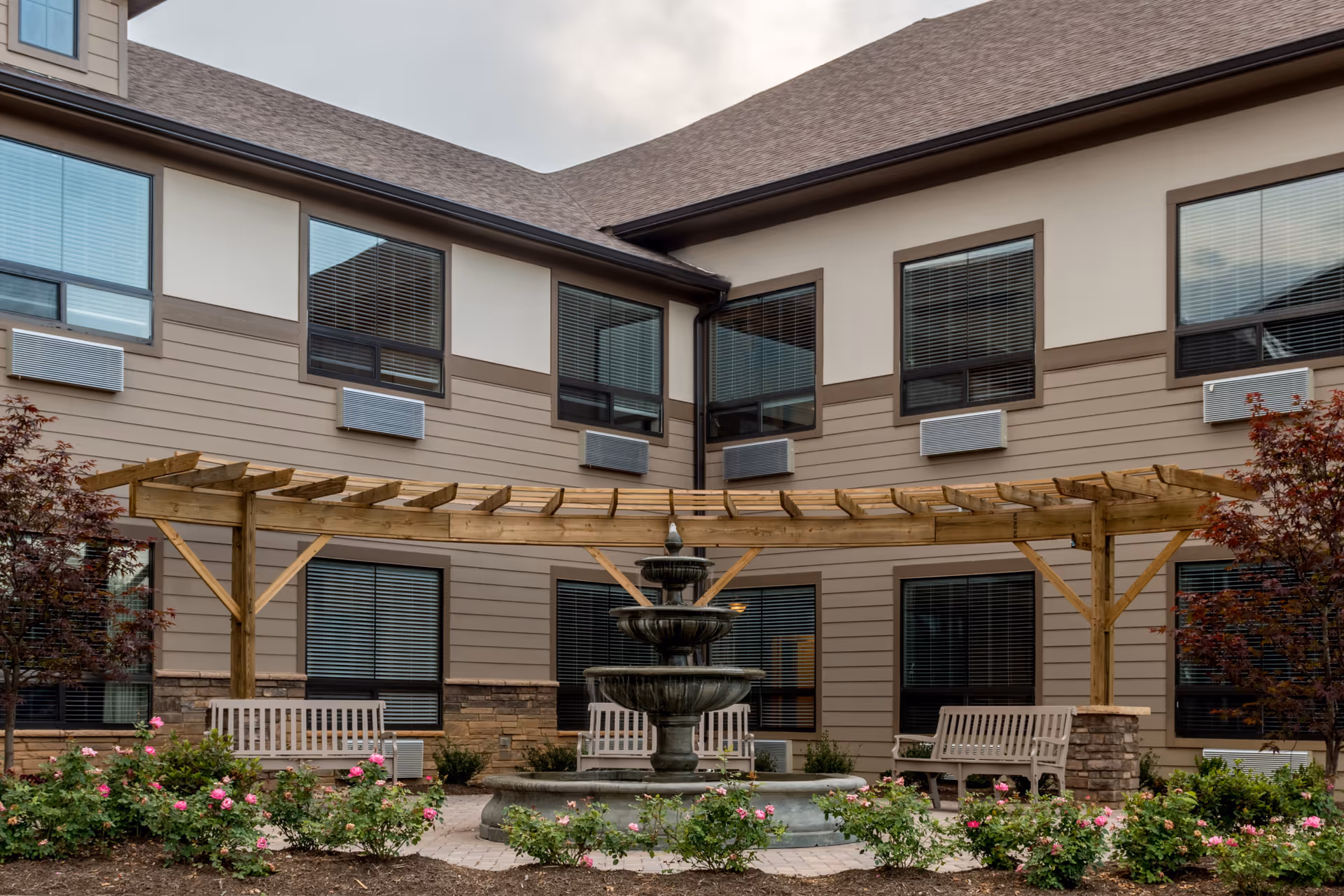 Outdoor courtyard area of a senior living facility featuring a three-tiered water fountain in the center, surrounded by blooming pink rose bushes and greenery. There are three wooden benches placed under a wooden pergola structure. The building exterior has beige siding with brown trim and multiple windows with blinds.