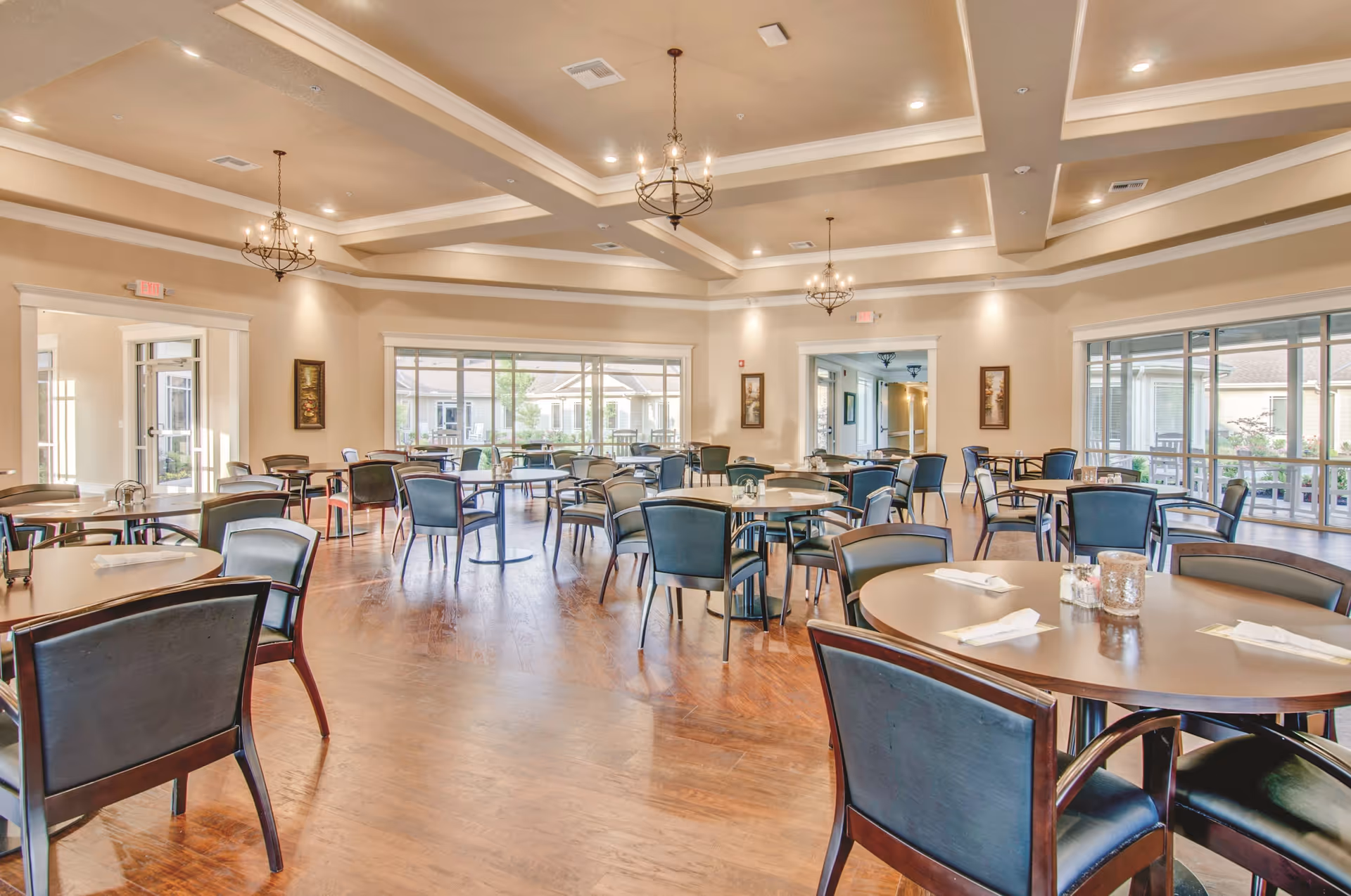 A spacious dining room with multiple round tables and black chairs arranged neatly. The room has large windows allowing natural light to fill the space, wooden flooring, and elegant chandeliers hanging from a coffered ceiling. The walls are painted in a light beige color with framed artwork.