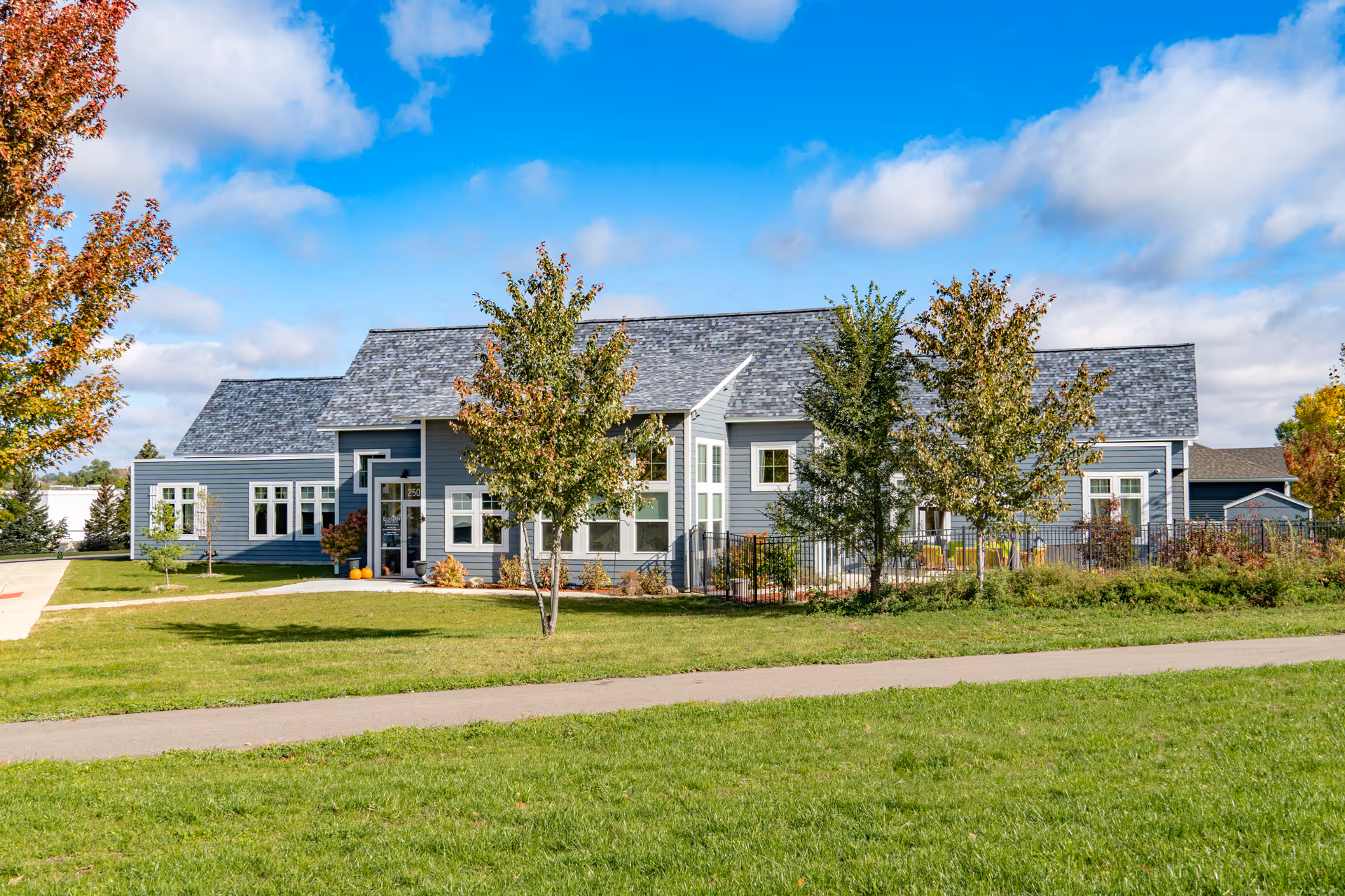Front exterior of a single-story blue senior living building with a lawn, trees, and a paved walkway under a partly cloudy sky.