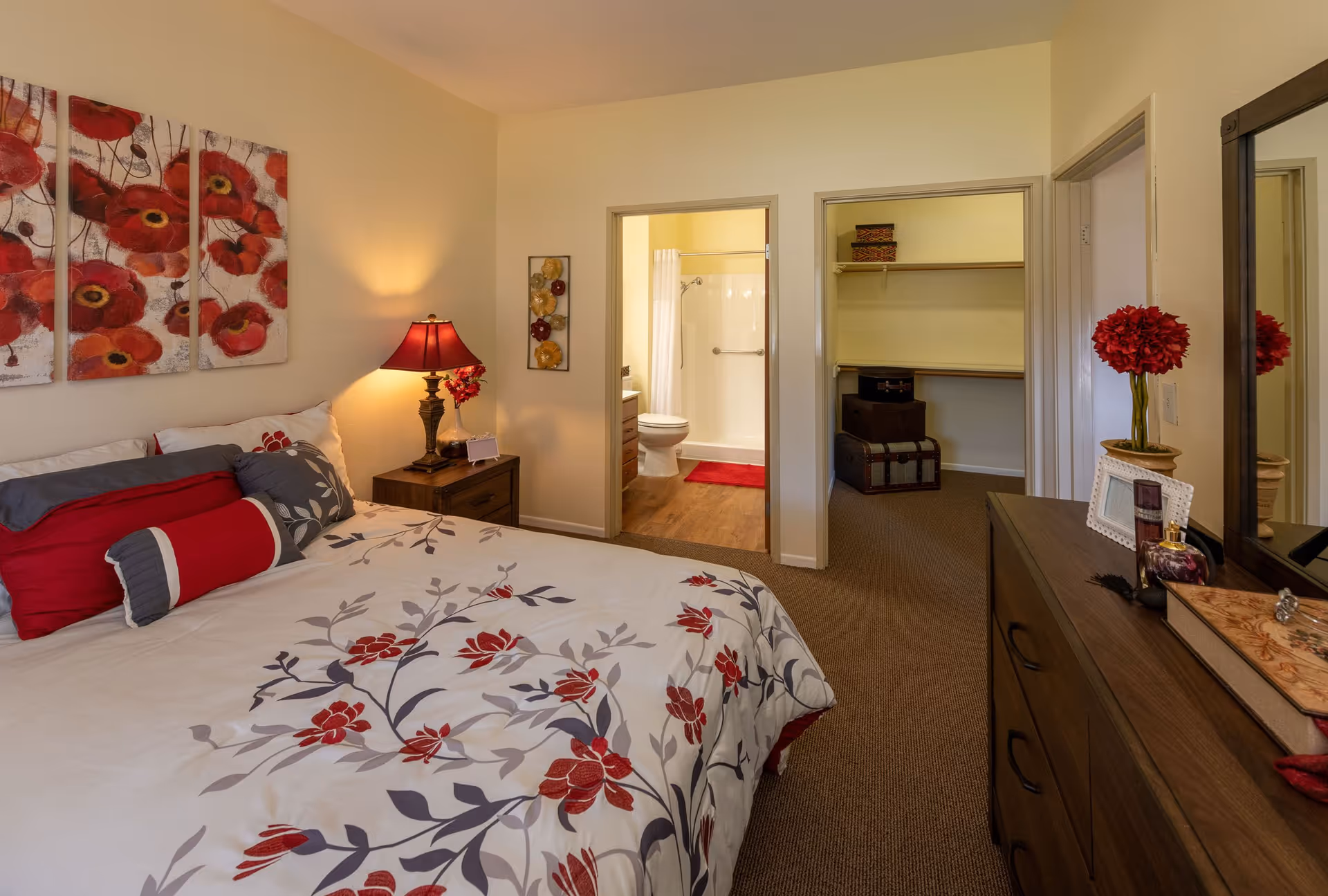 A cozy bedroom with a bed covered in white bedding with red and gray floral patterns. There is a nightstand with a lamp and flowers next to the bed. On the wall above the bed are three panels of red flower artwork. The room has two doorways; one leads to a bathroom with a shower, toilet, and red bath mat, and the other leads to a walk-in closet with shelves and storage boxes. A dresser with a mirror, decorative items, and a vase with red flowers is on the right side of the room.