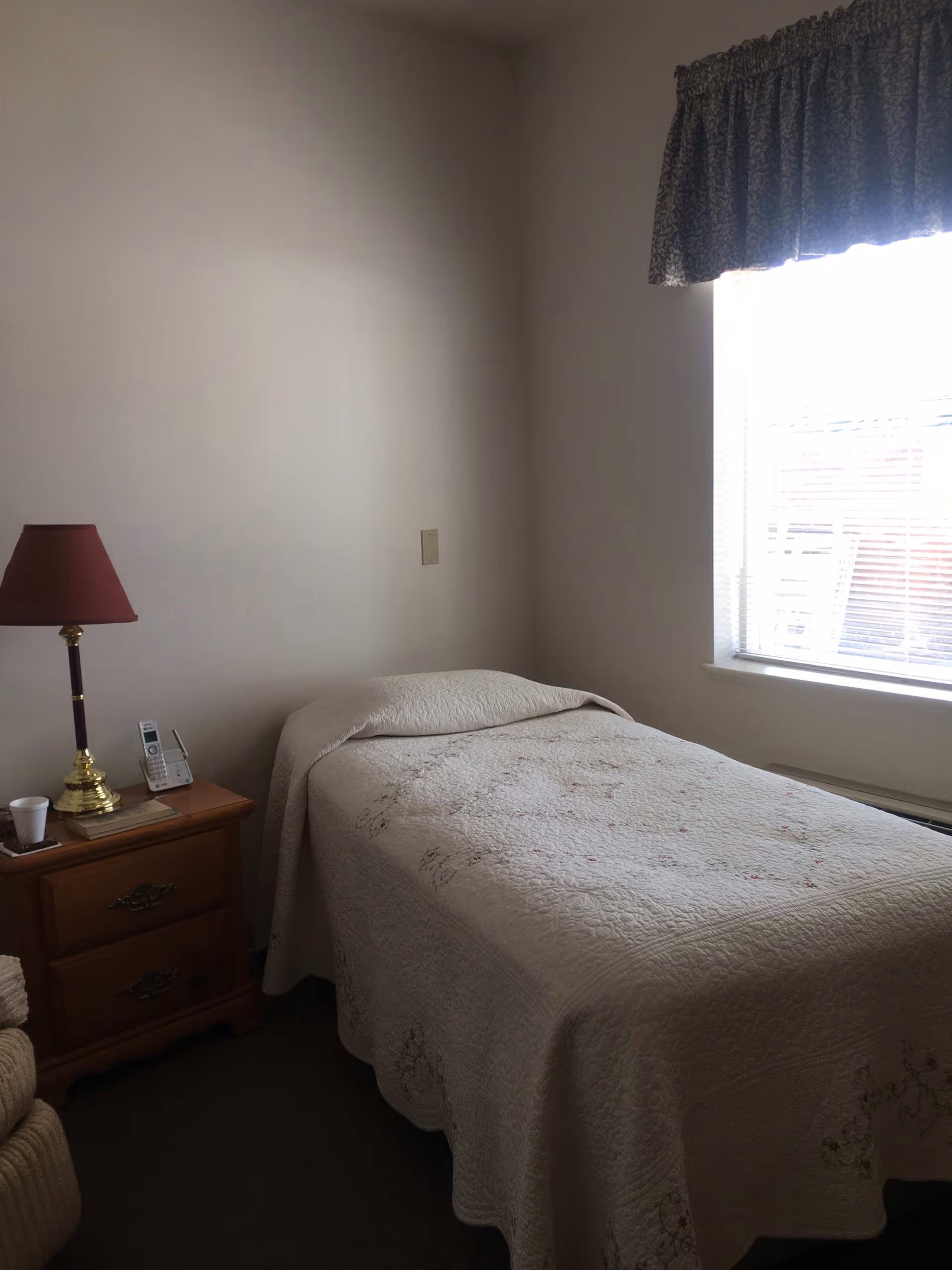 A small bedroom with a single bed covered with a white quilted bedspread. Next to the bed is a wooden nightstand with two drawers, a red lampshade on a brass lamp, a cordless phone, a book, and a white cup. A window with blinds and a patterned valance lets in natural light.