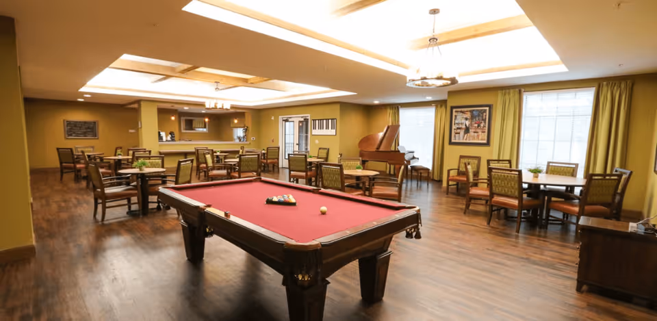 A spacious common area with a red felt pool table in the center, surrounded by multiple round tables and chairs. The room features wooden flooring, green walls, large windows with green curtains, and a grand piano in the corner. The ceiling has recessed lighting and decorative light fixtures.