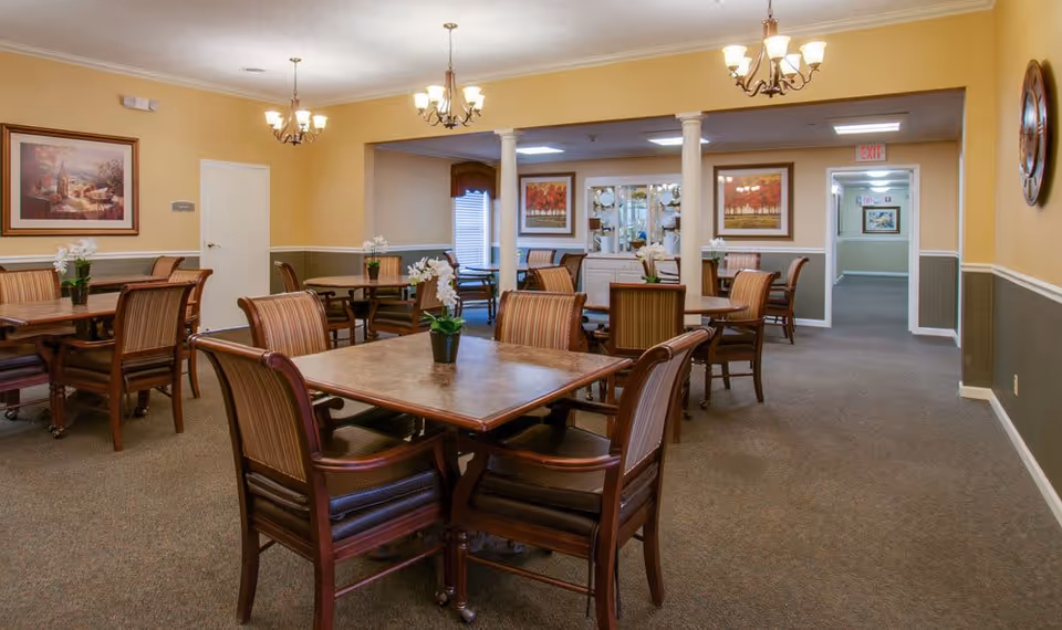 Communal dining room with several wooden tables and upholstered chairs, chandeliers, and framed artwork on warm yellow walls.