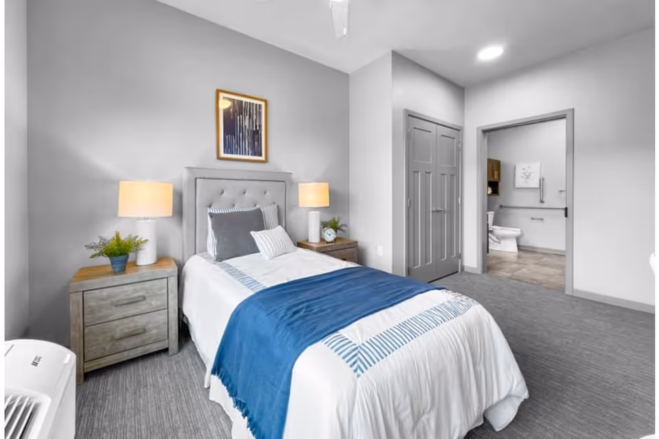 A neatly made single bed with white and blue bedding in a bedroom. There are two wooden nightstands on either side of the bed, each with a lamp and a small plant. A framed abstract artwork hangs above the headboard. The room has gray walls and carpeted flooring. An open doorway reveals a bathroom with a toilet and grab bars.
