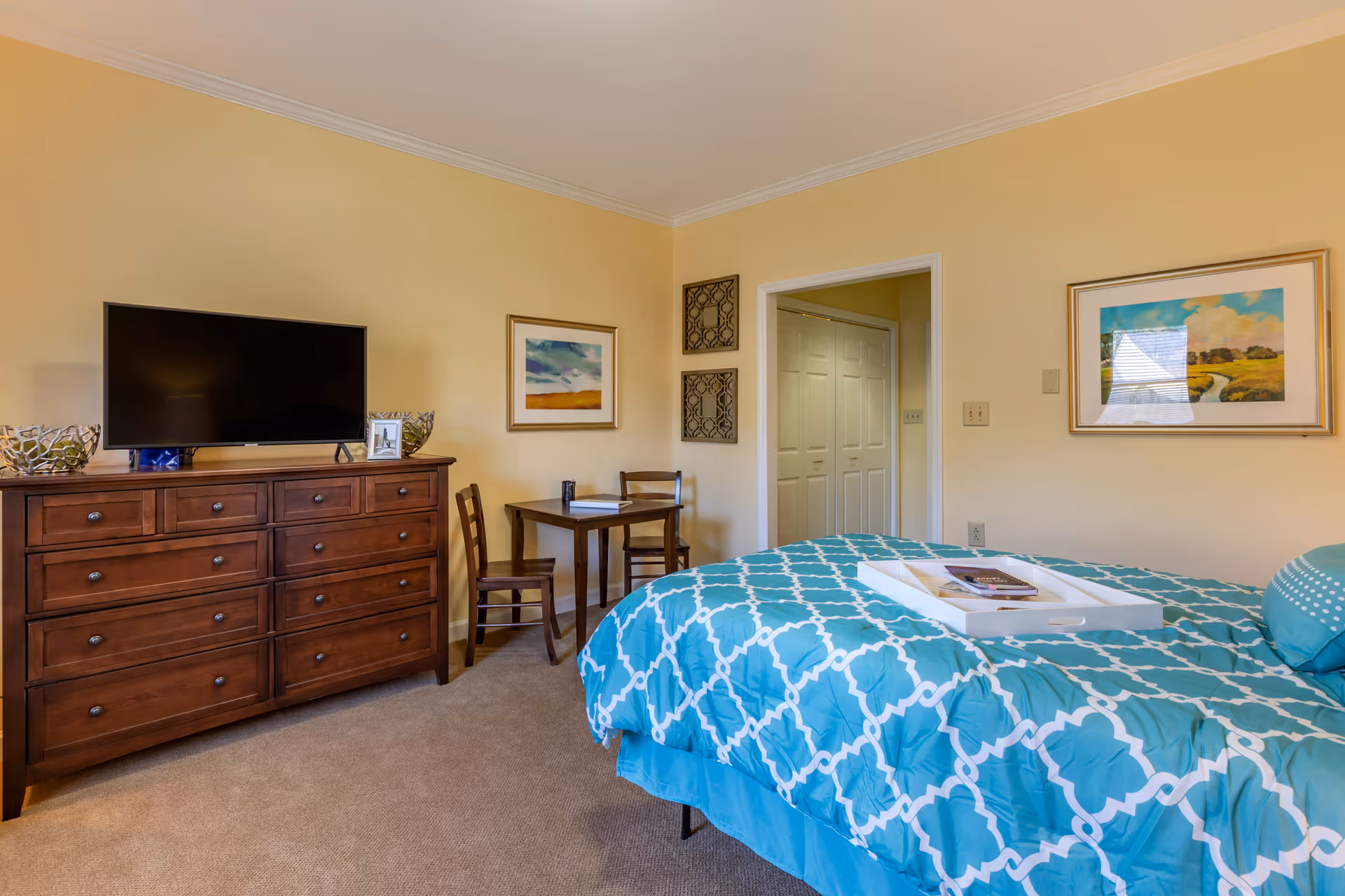 A bedroom with beige walls and carpeted floor featuring a bed with a blue and white patterned comforter. A white tray with books is placed on the bed. Against one wall is a wooden dresser with a flat-screen TV on top, flanked by decorative bowls. A small wooden table with two chairs is positioned in the corner near a doorway with white double doors. Two framed paintings and two decorative wall hangings adorn the walls.