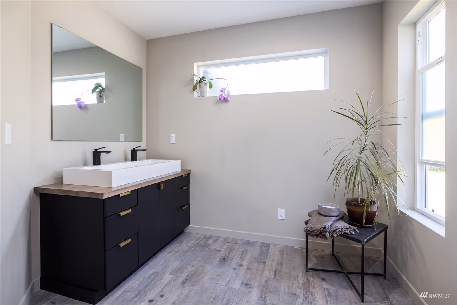 A modern bathroom with a large rectangular white sink on a black cabinet with brass handles, a large mirror above the sink, a small rectangular window with a potted orchid, and a corner table holding a potted plant, a folded towel, and a bowl. The floor has wood-like tiles and the walls are light-colored.