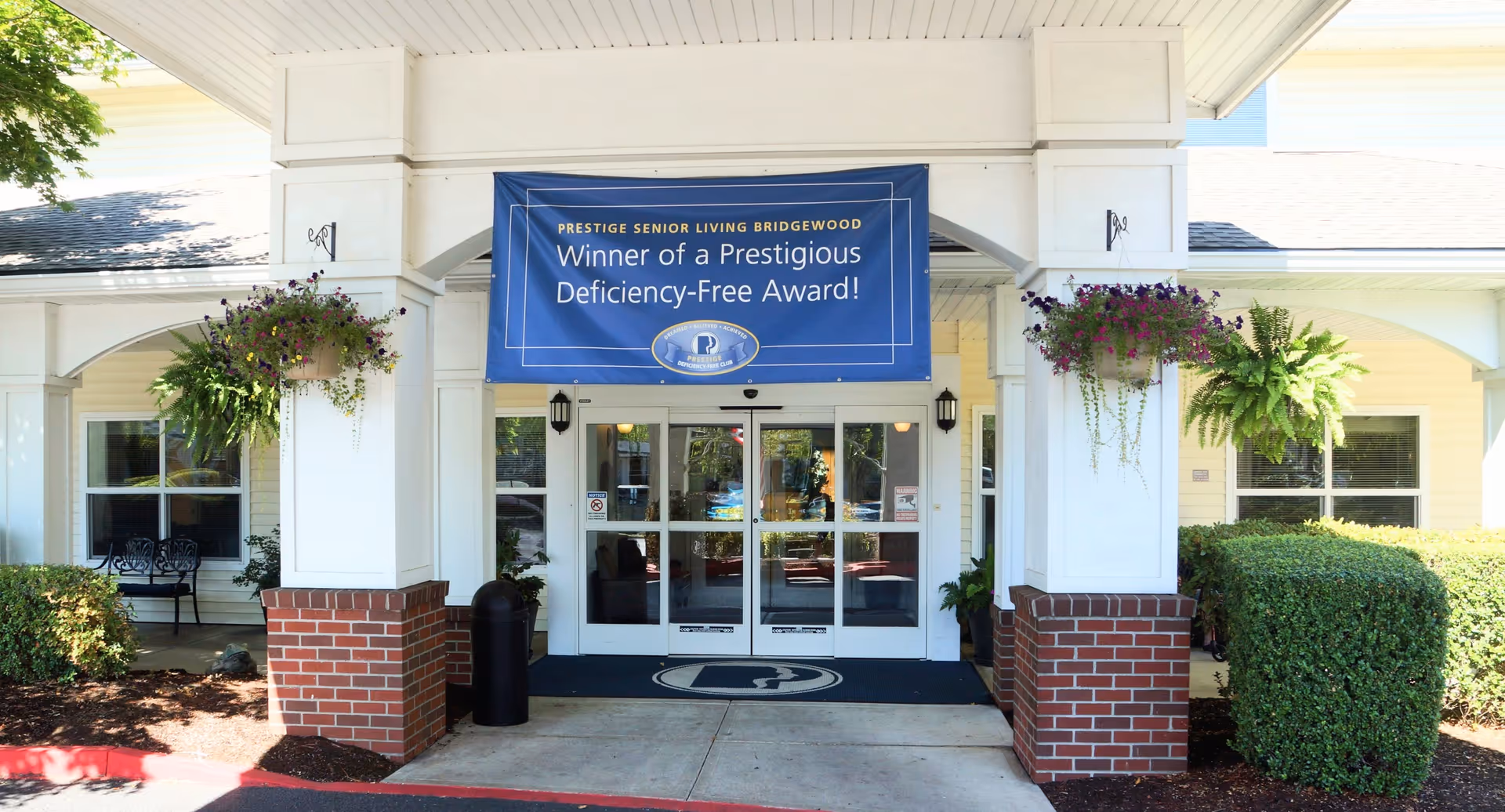 Entrance of Prestige Senior Living Bridgewood with a blue award banner above glass doors, brick columns, and hanging flower baskets.