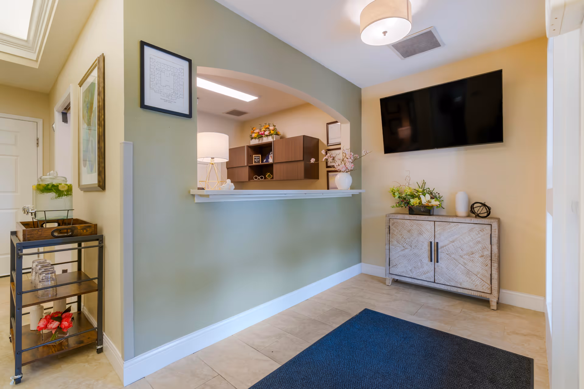Interior view of a reception area in a senior living facility with a green half-wall and an archway opening to an office space. A flat-screen TV is mounted on the wall above a wooden cabinet decorated with flowers and ornaments. A small cart with a glass beverage dispenser and glasses is positioned near the entrance door. The floor is tiled and a dark blue rug is placed in front of the cabinet.