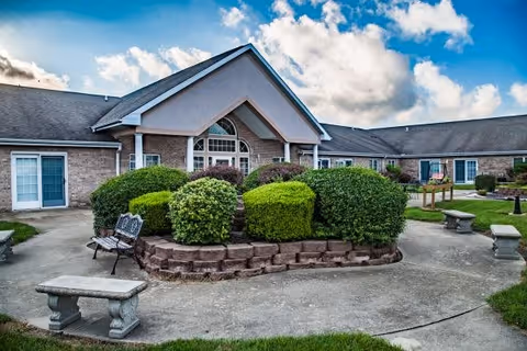 Outdoor courtyard area of a senior living facility with neatly trimmed bushes in the center, surrounded by a circular concrete pathway. There are stone benches and a metal bench around the courtyard. The building has a brick exterior with multiple windows and a peaked roof under a partly cloudy sky.