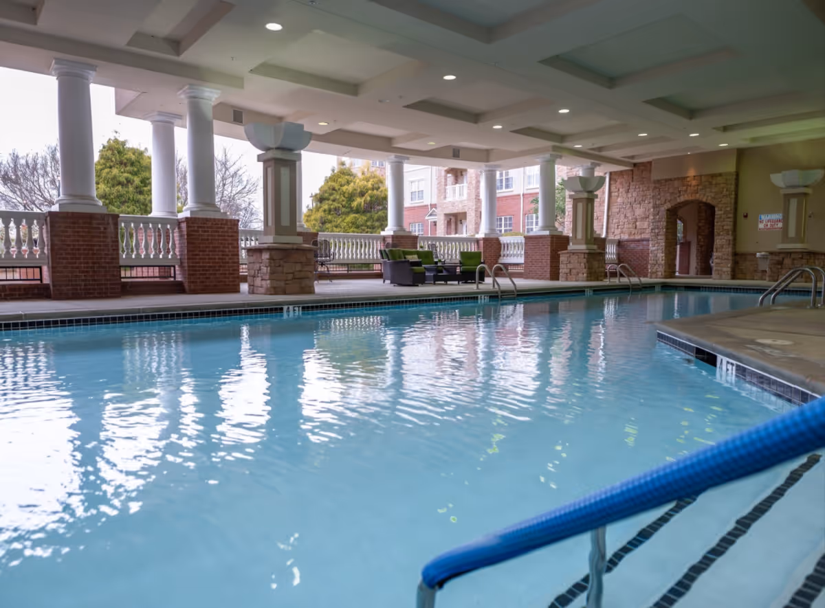 A covered indoor swimming pool with columns, lounge seating, and brick archways in a residential facility.