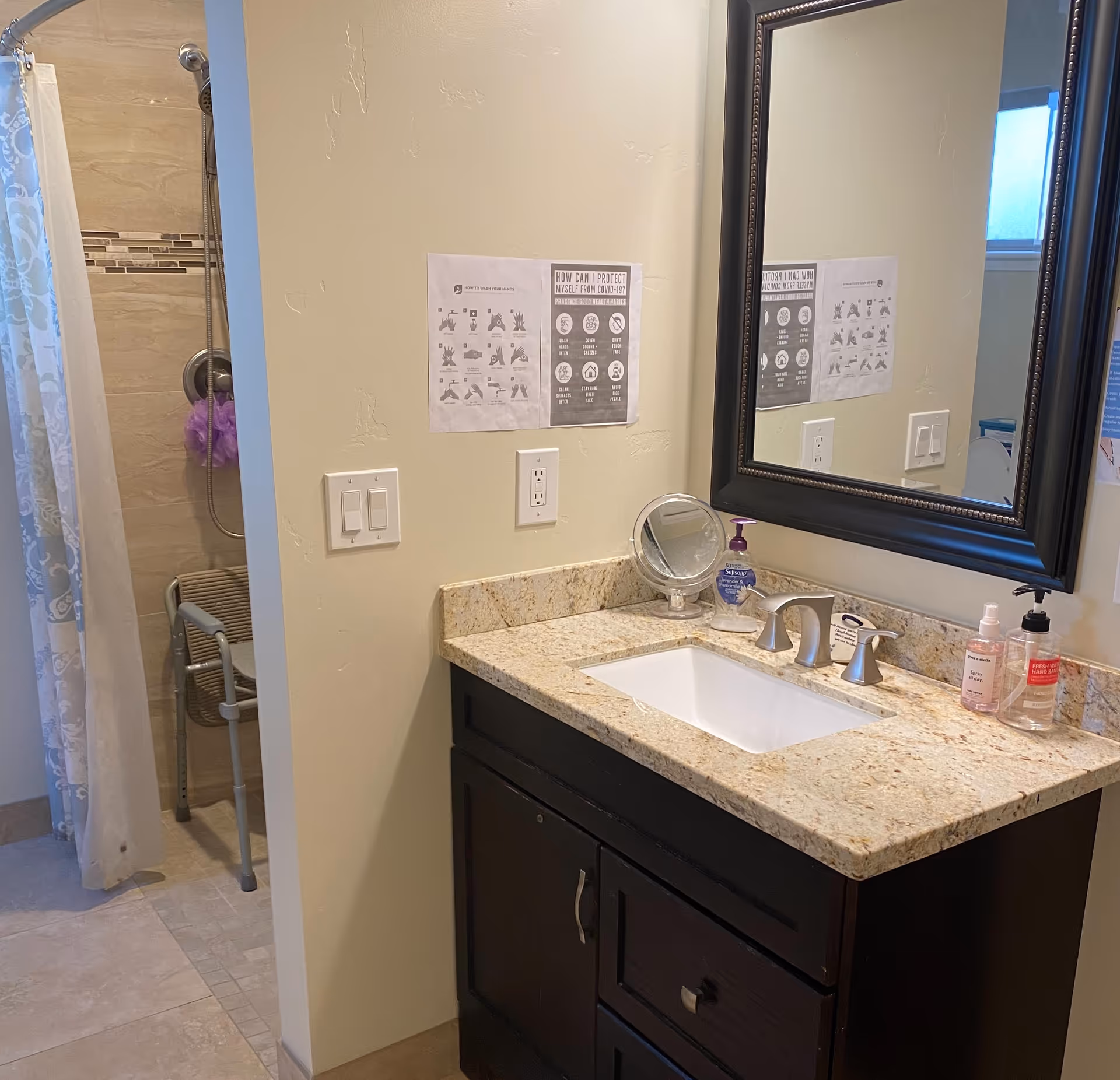Bathroom interior featuring a granite countertop with a sink, a large framed mirror above it, and various hand sanitizers and soap dispensers on the counter. To the left, there is a shower area with a curtain and a shower chair visible. Posters with COVID-19 protection and handwashing instructions are taped to the wall.