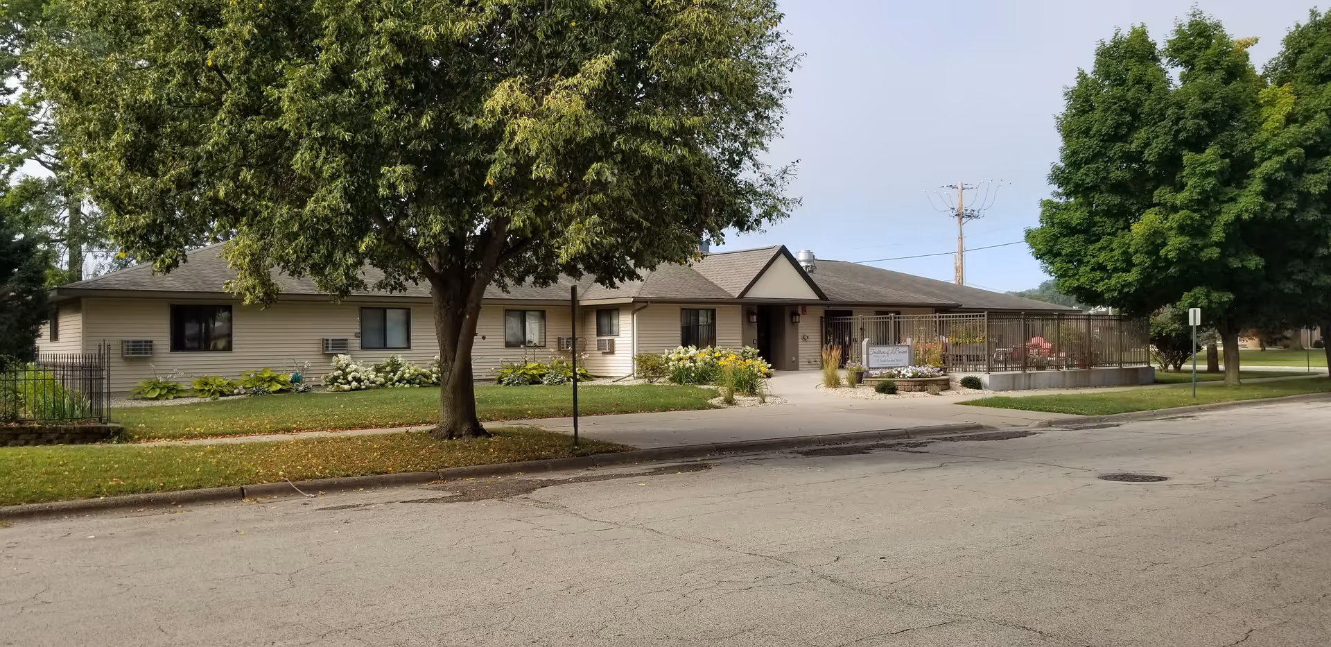 Front exterior of a single-story senior living facility with a driveway, fenced patio, landscaped grounds, and large trees.