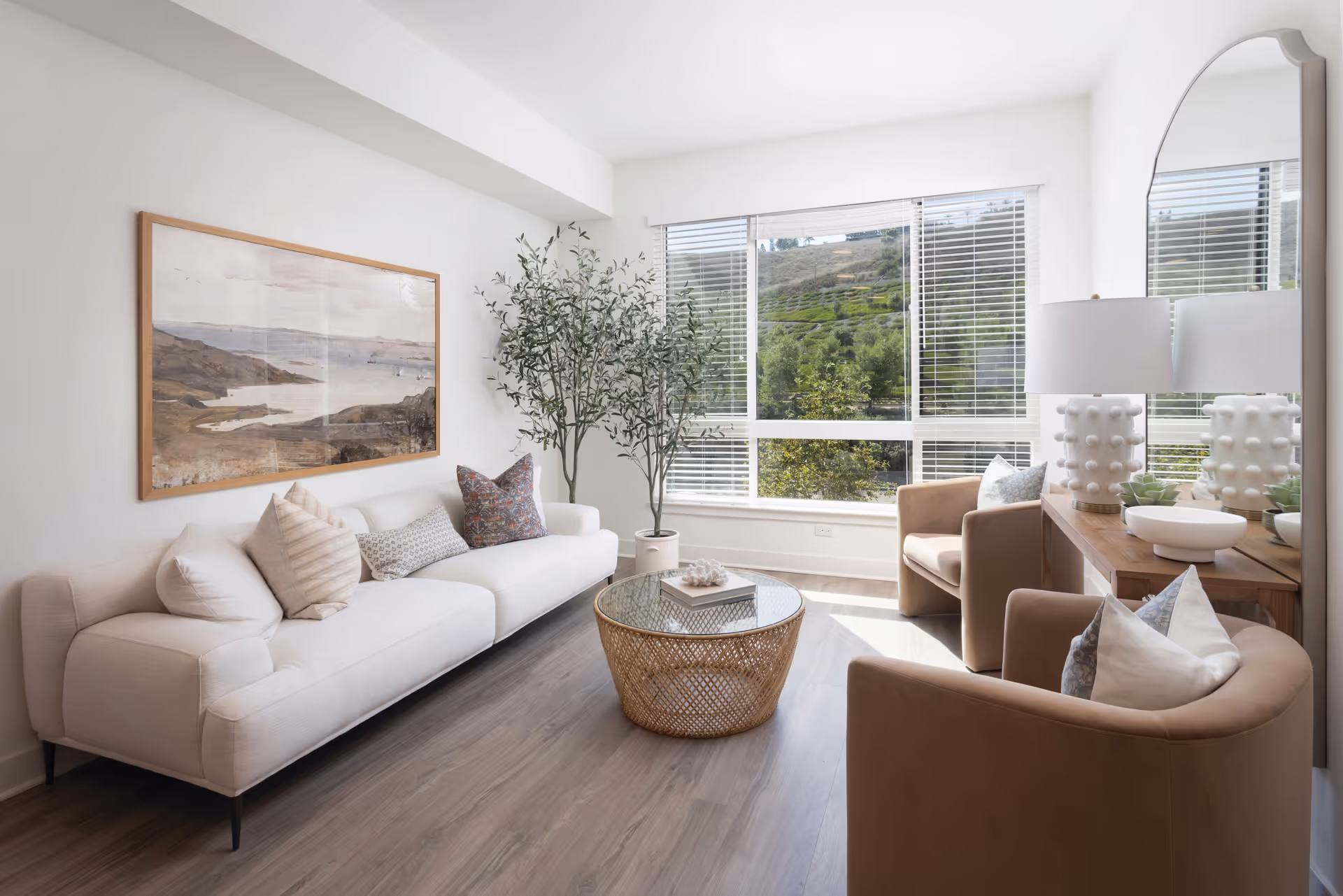 A bright and modern living room with a large window showing a green hillside outside. The room features a white sofa with multiple pillows, two beige armchairs with pillows, a round wicker coffee table with a glass top, a wooden console table with a white lamp and decorative items, a large wall mirror, a framed landscape painting on the wall, and a tall potted plant in the corner.