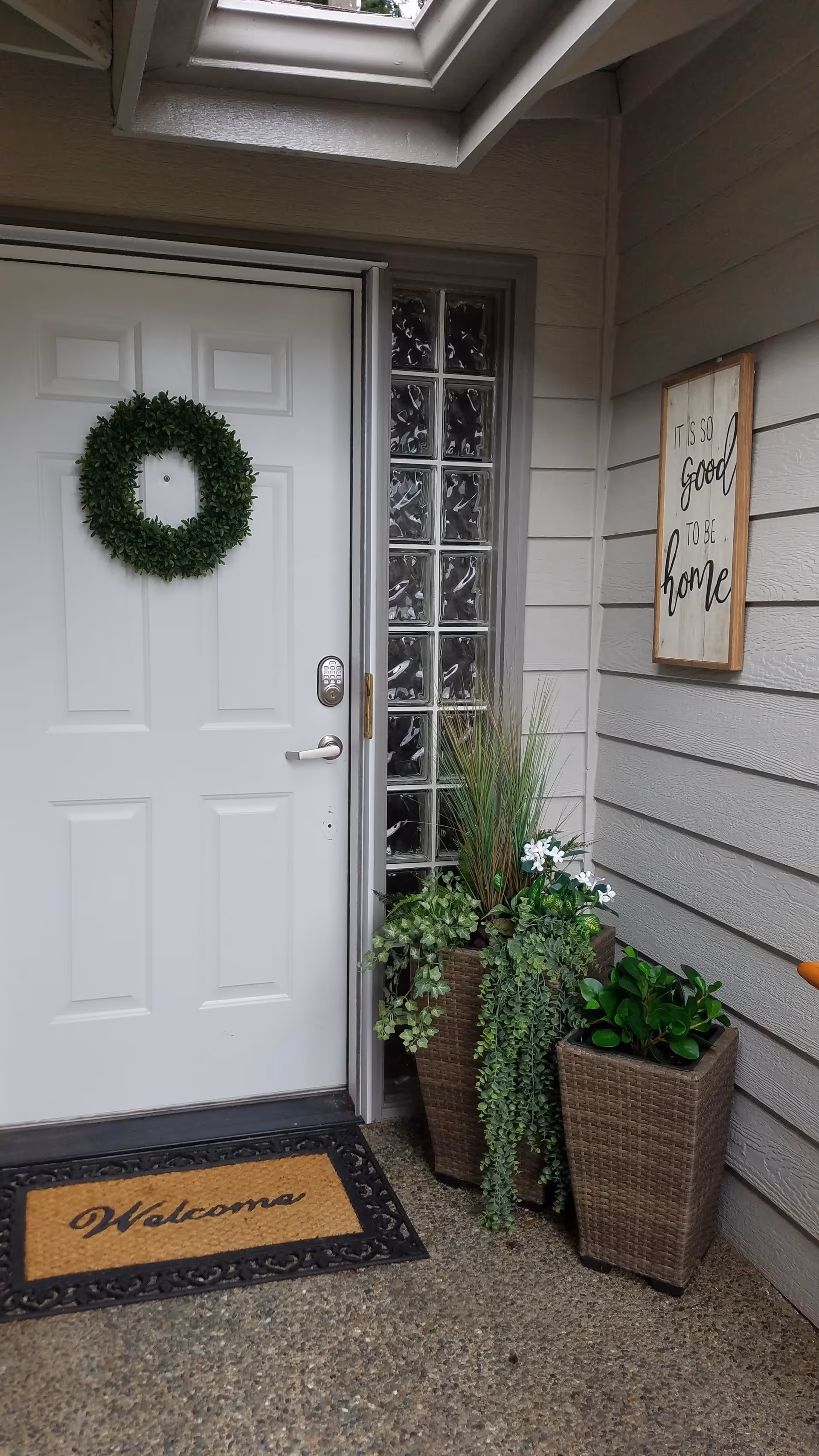 Front entrance of a facility with a white door decorated with a green wreath. There is a welcome mat on the ground and two tall planters with green plants next to the door. A sign on the wall reads 'It is so good to be home'.