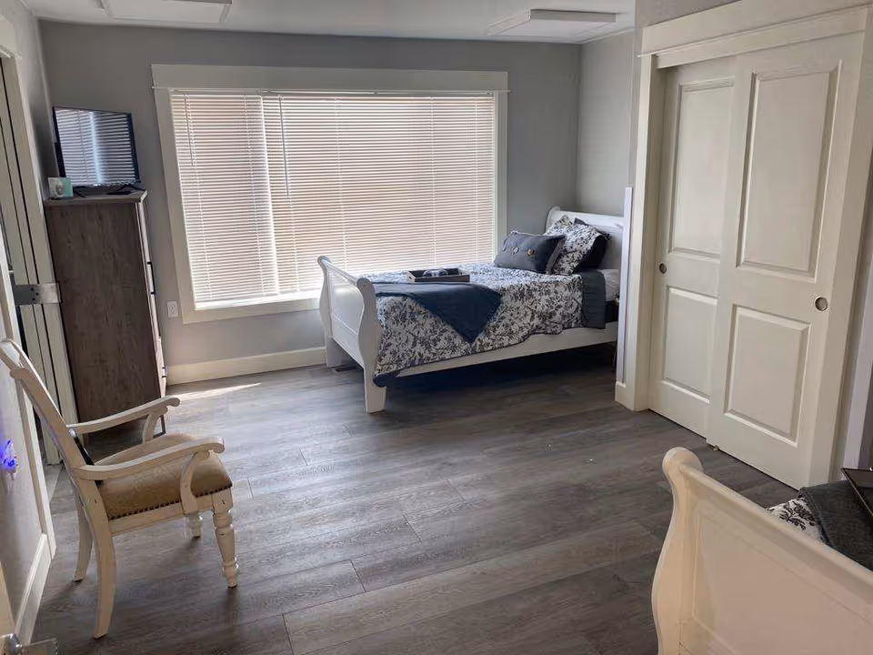 Bright, minimally furnished bedroom with a white sleigh bed by a large window, wooden flooring, dresser, and chairs.
