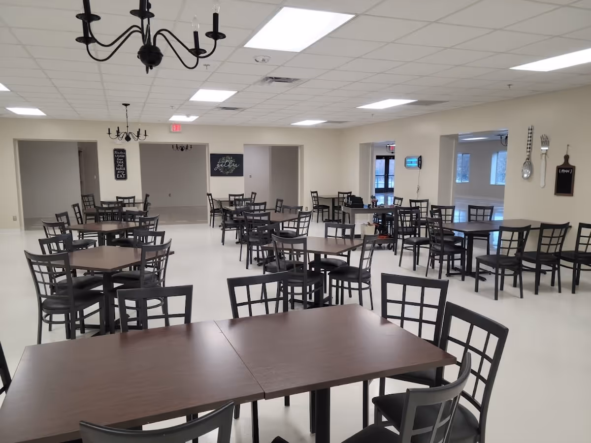 A spacious dining room with multiple dark wooden tables and black chairs arranged neatly. The room has a white floor and beige walls, with black chandeliers hanging from the ceiling. Wall decorations include a large spoon and fork, a small chalkboard, and framed signs with inspirational messages. Large windows and open doorways provide natural light and views into adjacent rooms.
