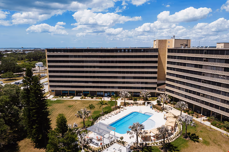 A large multi-story senior living facility building with a central outdoor swimming pool surrounded by lounge chairs and palm trees under a partly cloudy blue sky.