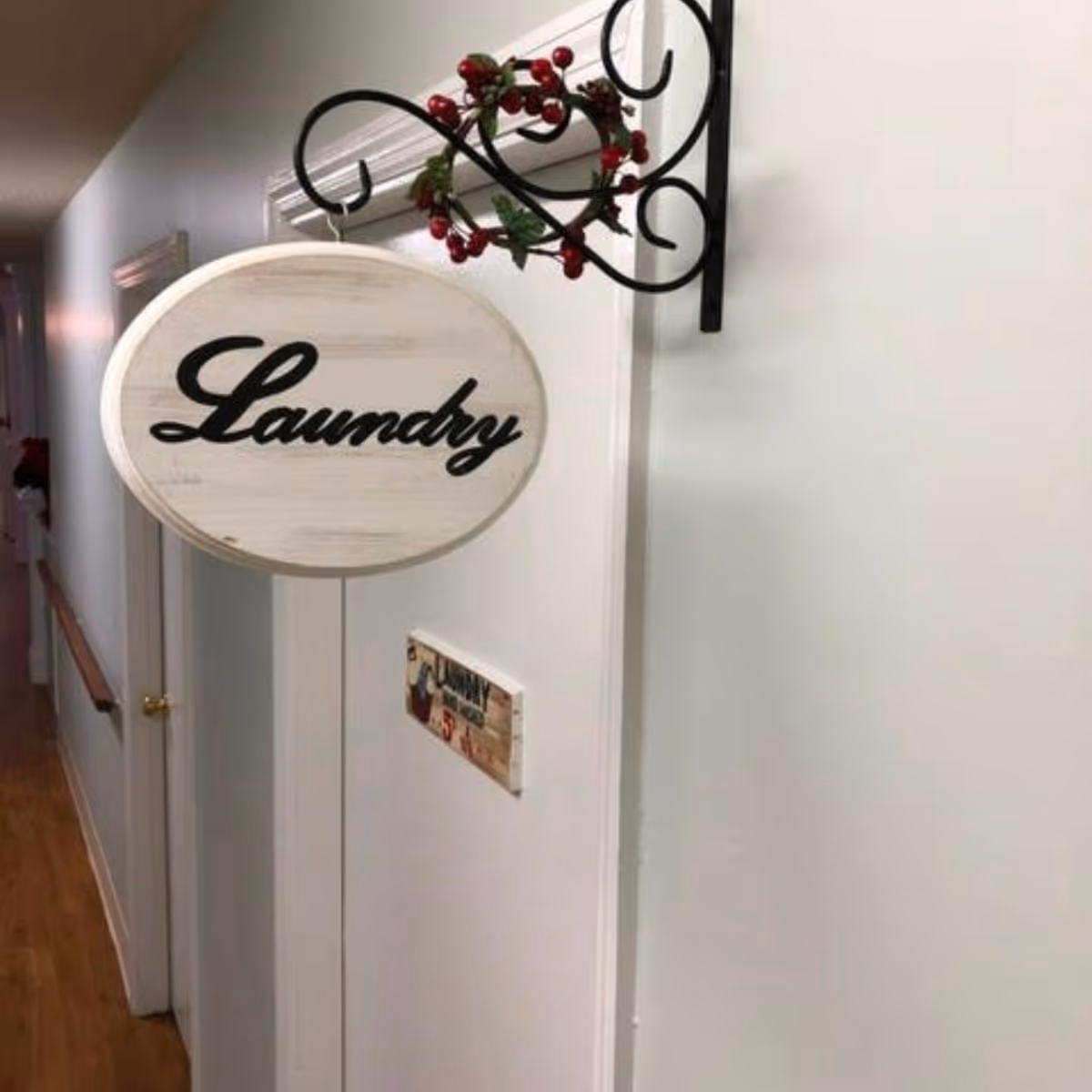 A white wooden door with a hanging oval sign that reads 'Laundry' in black cursive letters. The sign is attached to a decorative black metal bracket adorned with a small wreath of red berries and green leaves. The door is part of a hallway with light-colored walls and wooden flooring.