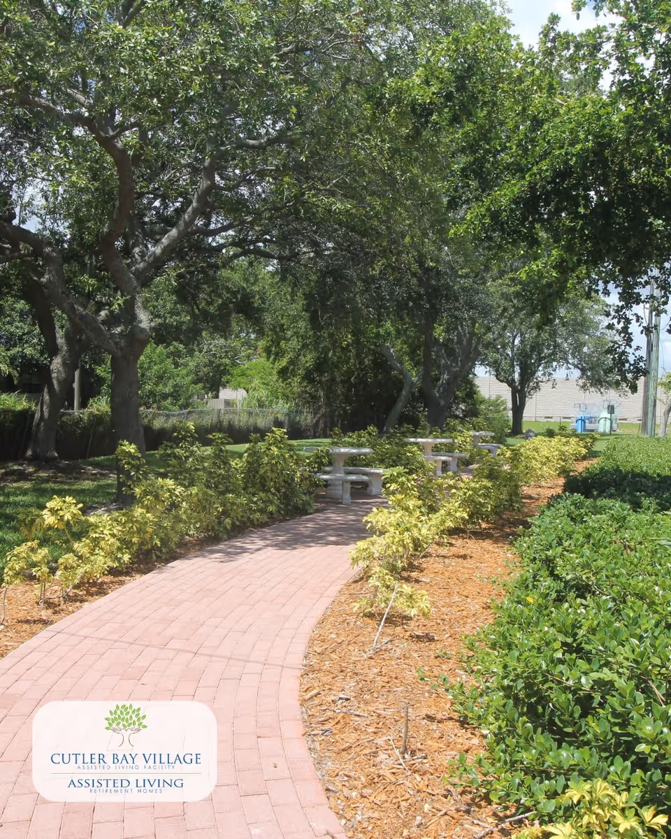 A paved brick pathway winding through a garden area with green shrubs and trees on both sides. Several concrete picnic tables and benches are visible along the path under the shade of large trees. The sky is partly cloudy, and the setting appears peaceful and well-maintained.