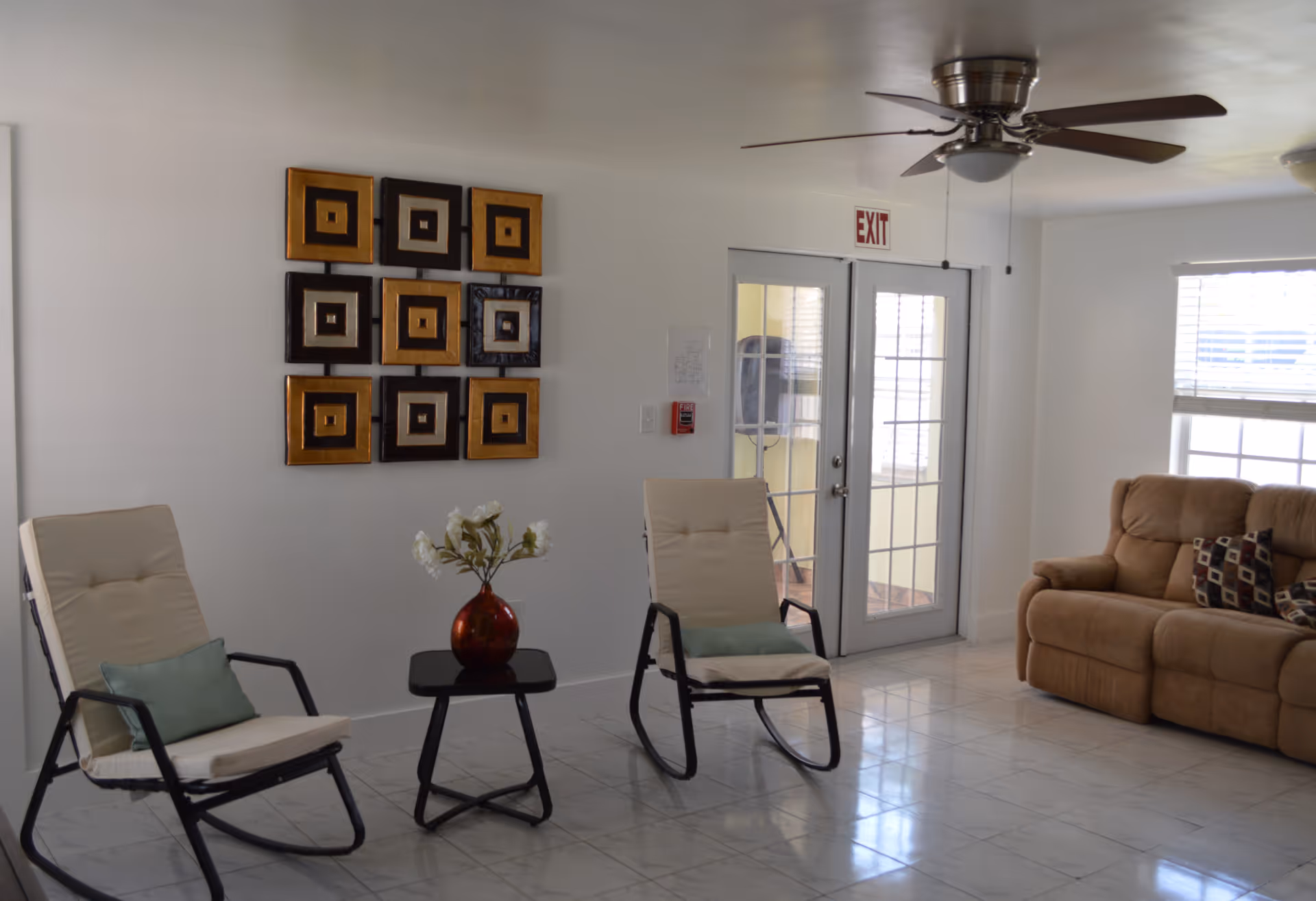 A bright living room with two cushioned chairs with light green pillows, a small black side table with a red vase holding white flowers, a brown sofa with patterned pillows, a ceiling fan, and a wall decorated with a grid of square art pieces. There is a glass door with an exit sign above it and a window with blinds letting in natural light.
