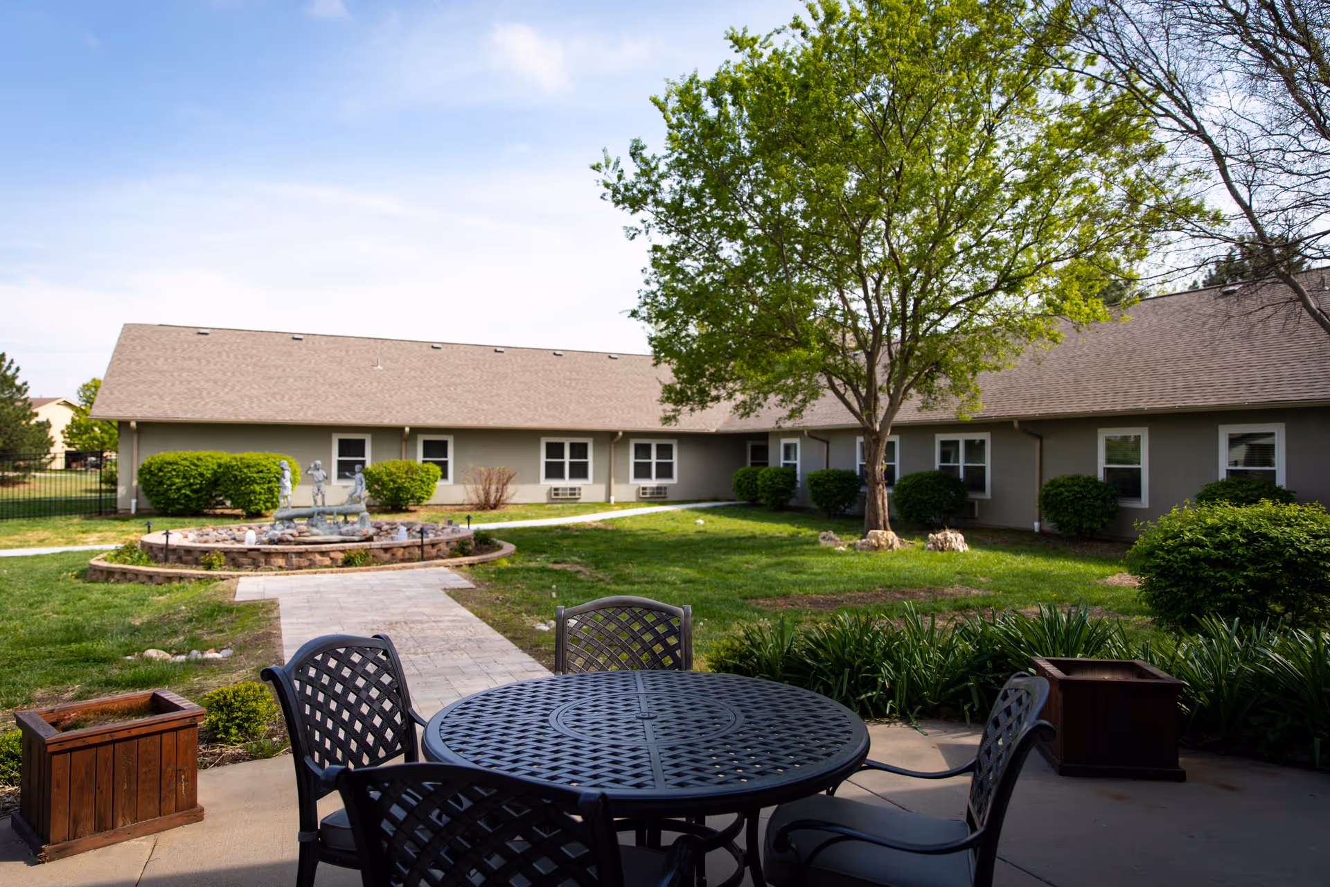 Outdoor courtyard with a metal patio table and chairs in the foreground, a central fountain, lawn, trees, and a single-story building in the background.