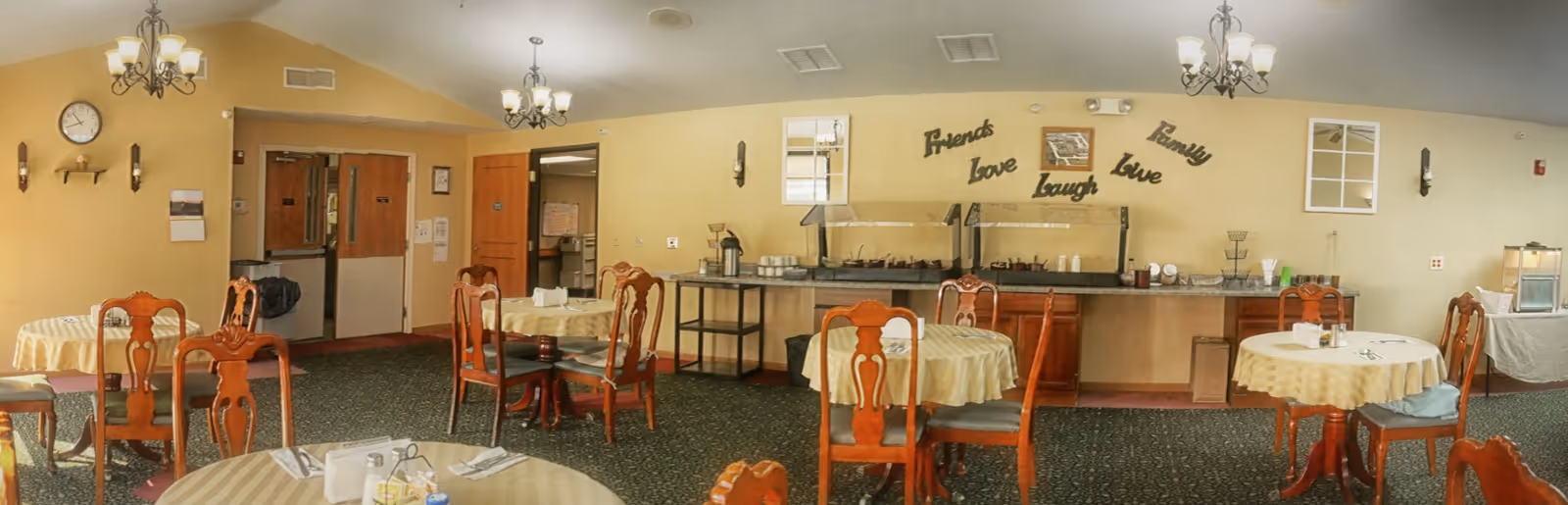 Dining room with round tables covered in beige tablecloths and wooden chairs. A buffet area with food warmers and coffee pots is against the wall. The wall has decorative words saying 'Friends', 'Family', 'Love', 'Laugh', and 'Live'. There are chandeliers hanging from the ceiling and a clock on the wall near double doors.