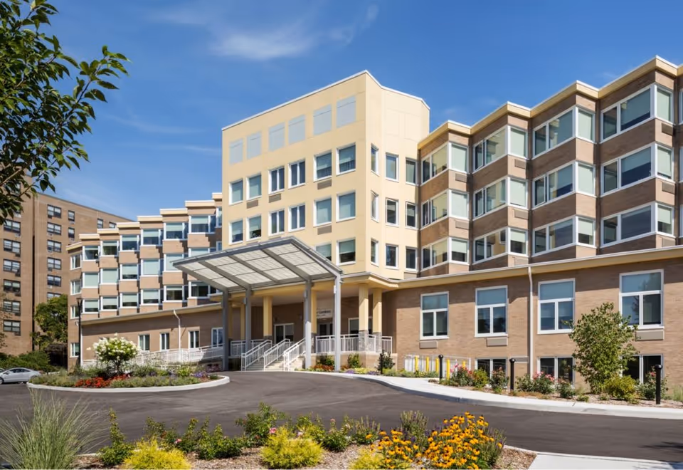 Front exterior of a multi-story memory care building with a covered entrance canopy, driveway and landscaped flower beds.